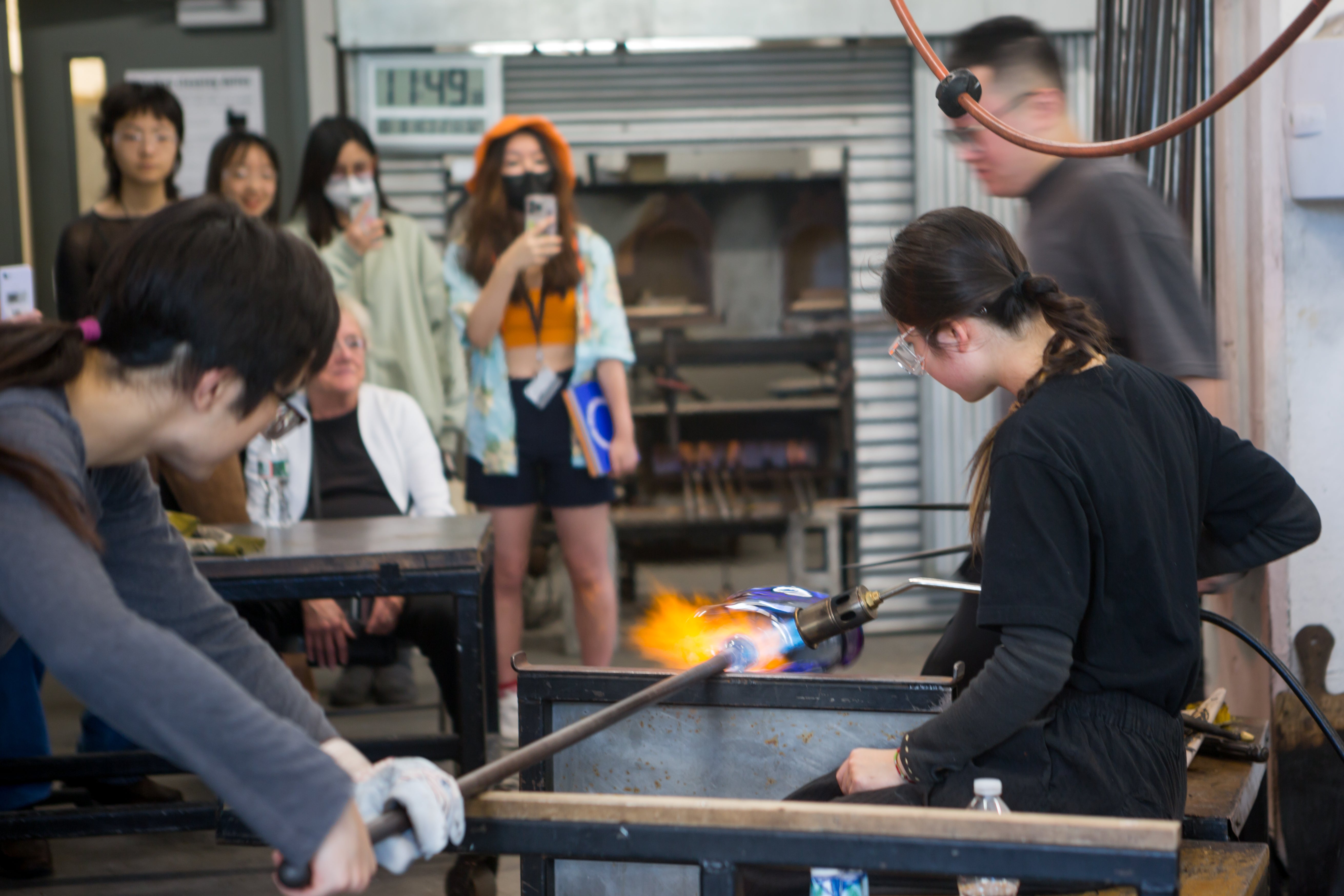 /Two%20students%20demonstrate%20blowing%20glass%2C%20using%20a%20torch%20to%20heat%20glass%20on%20a%20long%20pipe%2C%20%20while%20others%20look%20on%2C%20recording%20with%20their%20cellphones.