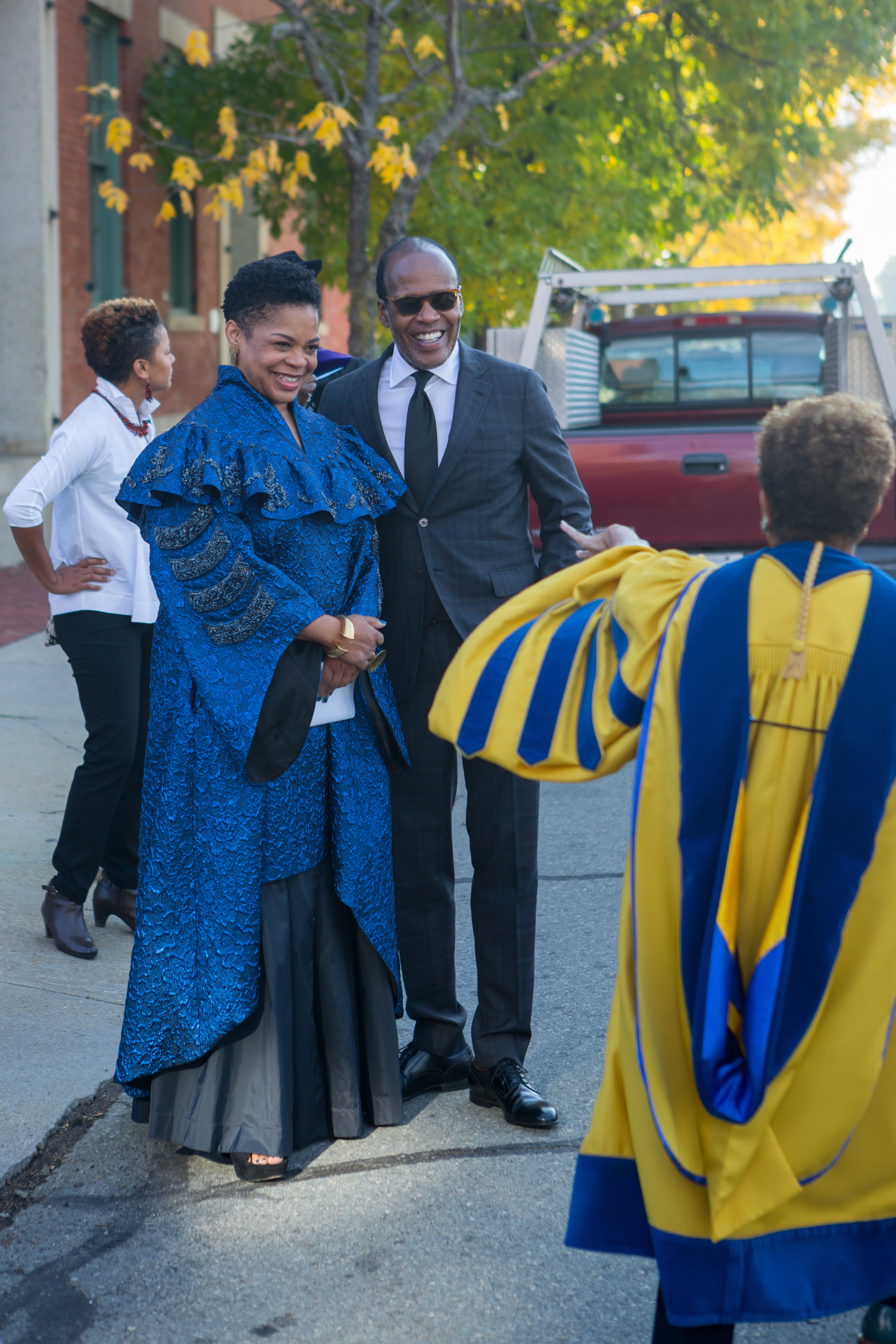 /A%20Black%20woman%20in%20blue%20ceremonial%20robes%20and%20a%20Black%20man%20in%20a%20suit%20greet%20a%20woman%20in%20yellow%20and%20blue%20doctoral%20robes