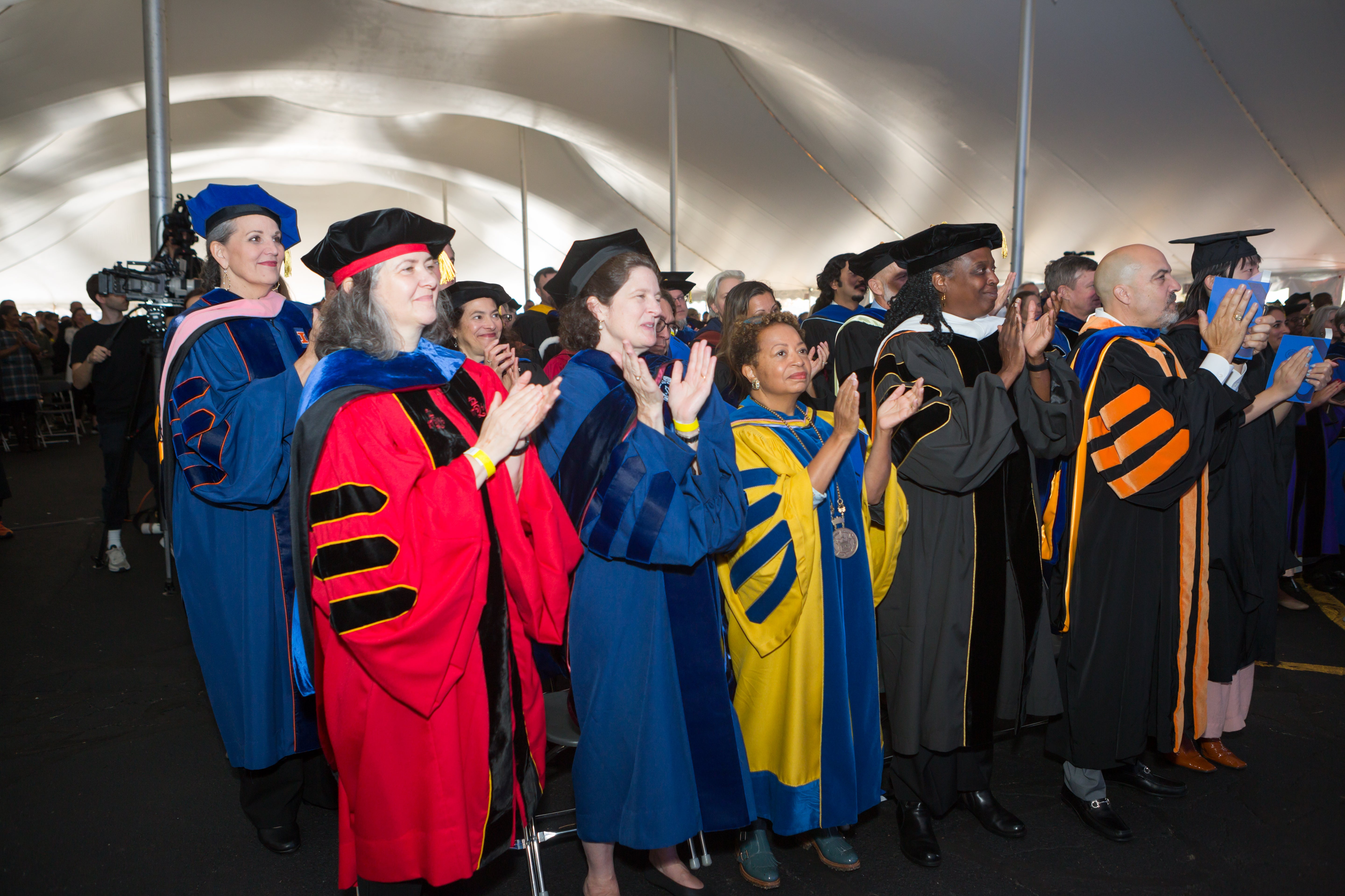 /A%20group%20of%20adults%20in%20doctoral%20robes%20of%20various%20colors%20stand%20and%20clap%20in%20a%20tent