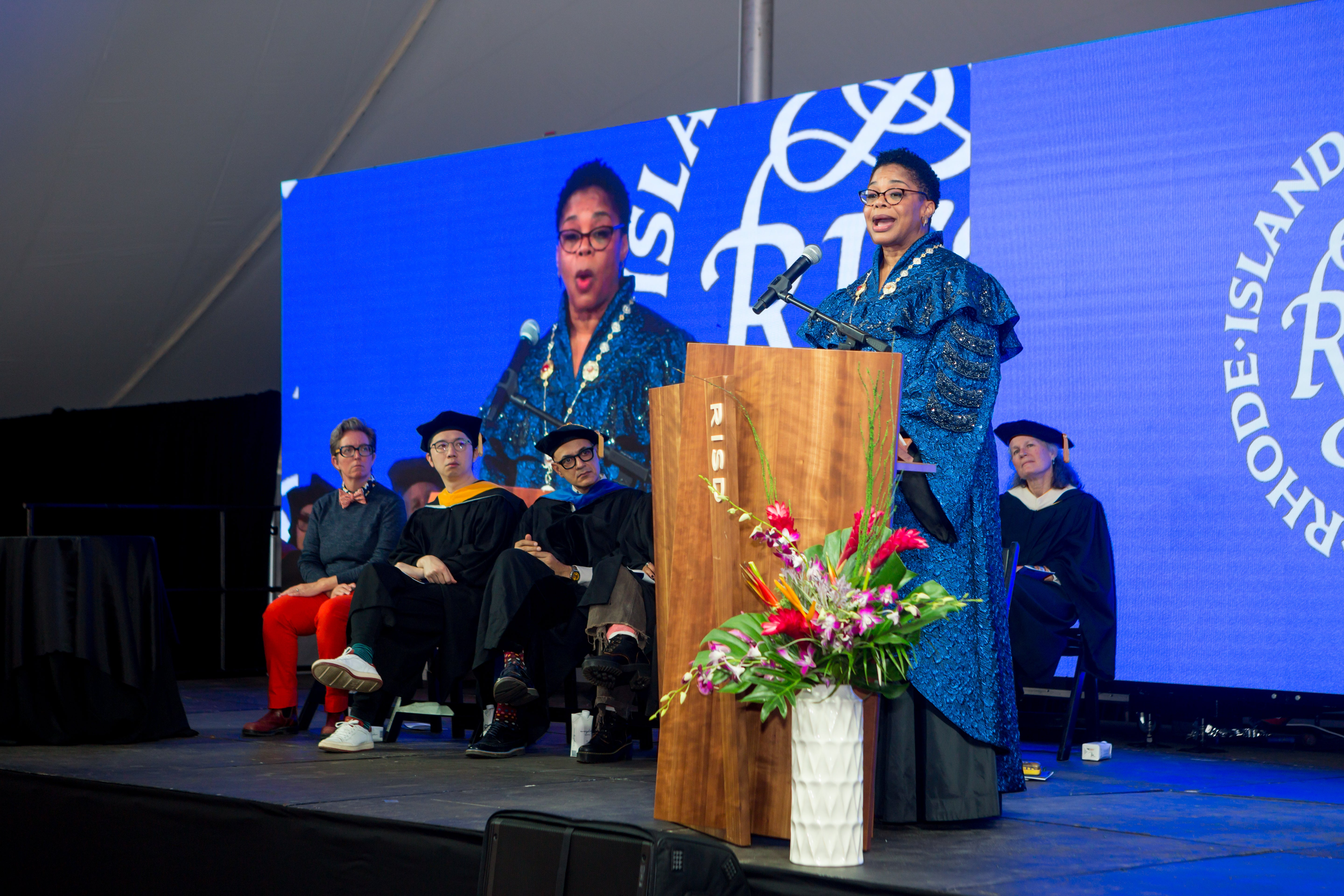/A%20Black%20woman%20in%20blue%20ceremonial%20robes%20speaks%20at%20a%20podium%2C%20on%20a%20stage%20where%20a%20group%20of%20onlookers%20sit.%20Her%20image%20is%20projected%20behind%20them.