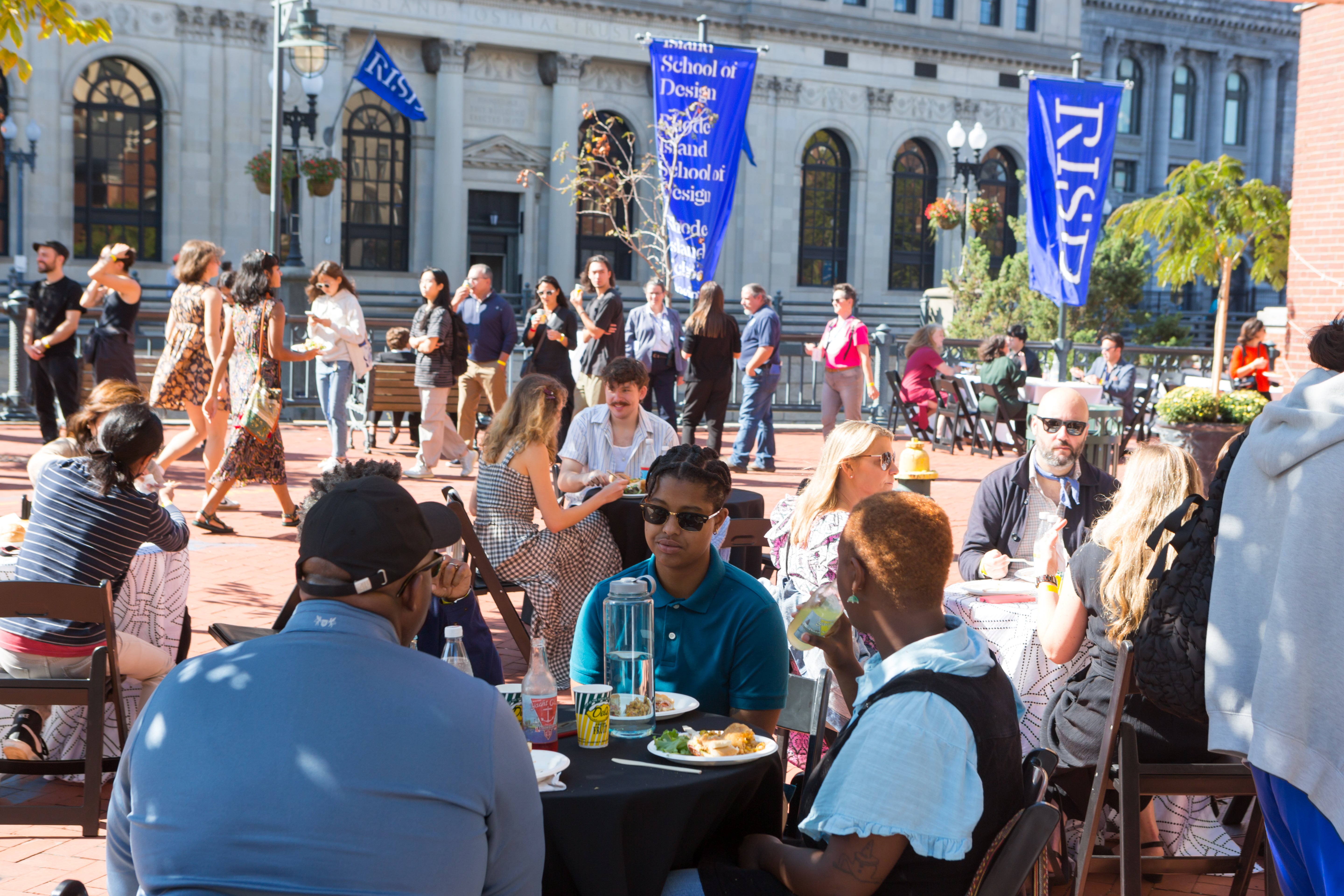 /On%20a%20sunny%20Fall%20day%2C%20groups%20sit%20and%20eat%20at%20small%20round%20tables.%20In%20the%20background%2C%20people%20walk%20along%20the%20Providence%20Riverwalk.%20Blue%20RISD%20banners%20hang%20from%20light%20poles.