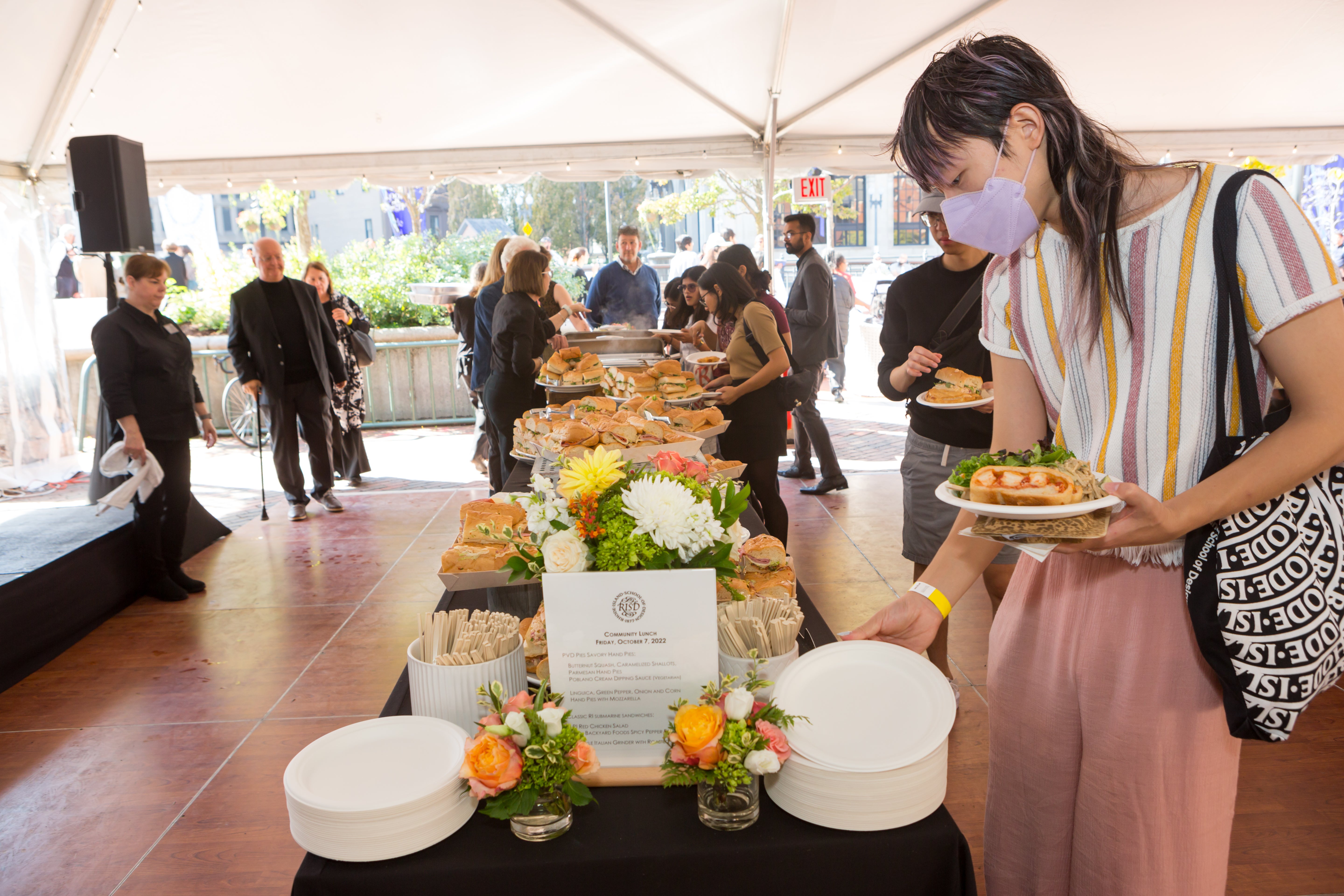 /Buffet%20line%20outdoors%20under%20a%20tent.%20A%20table%20is%20piled%20with%20sandwiches%2C%20plates%2C%20and%20flowers.%20A%20young%20AAPI%20person%20picks%20up%20a%20plate%20in%20the%20foreground%2C%20while%20a%20small%20group%20examines%20food%20offerings%20in%20the%20background.