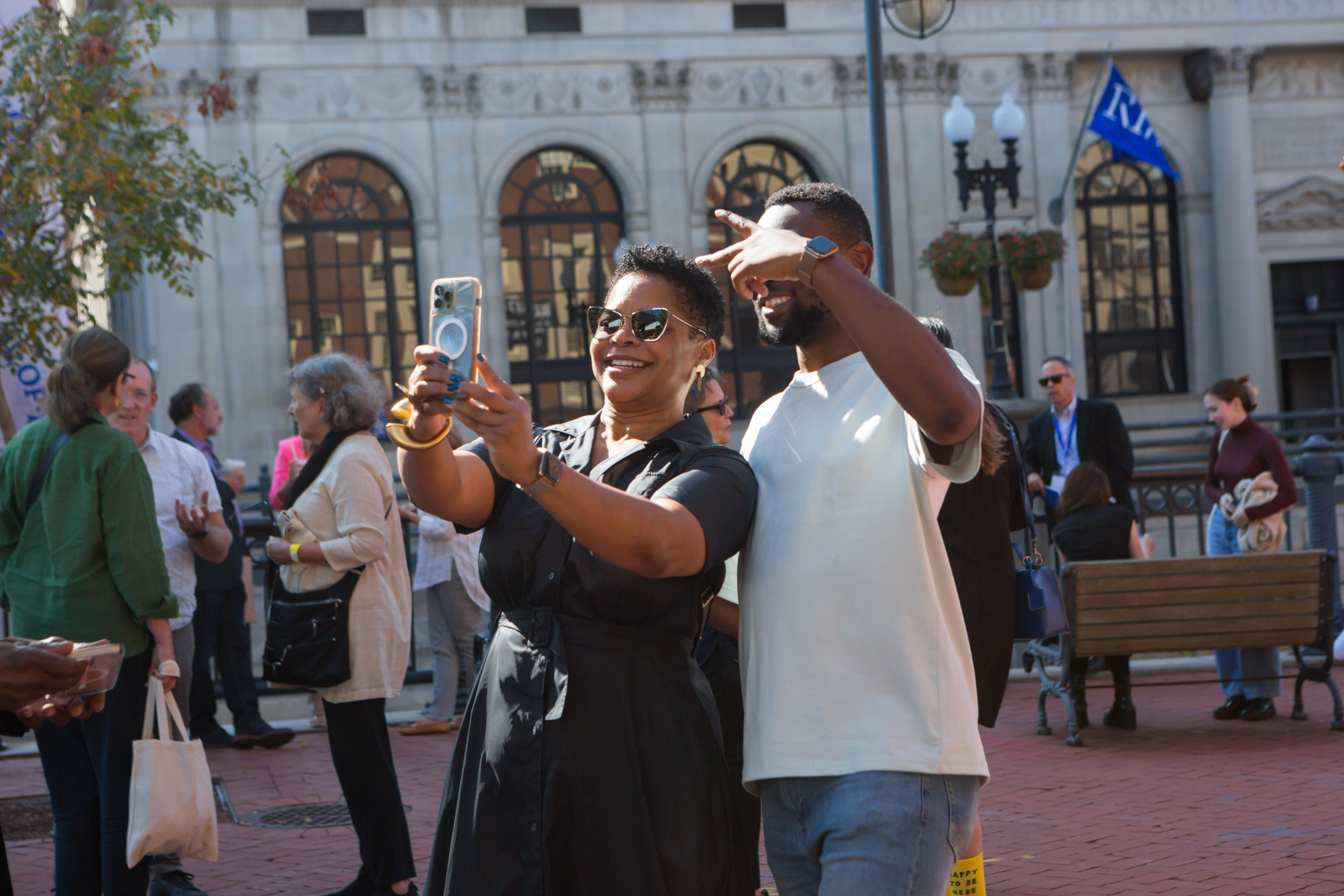 /Outside%20on%20a%20brick%20walkway%20a%20Black%20woman%20in%20sunglasses%20takes%20a%20selfie%20with%20a%20young%20black%20man%20in%20a%20white%20tee%20shirt.