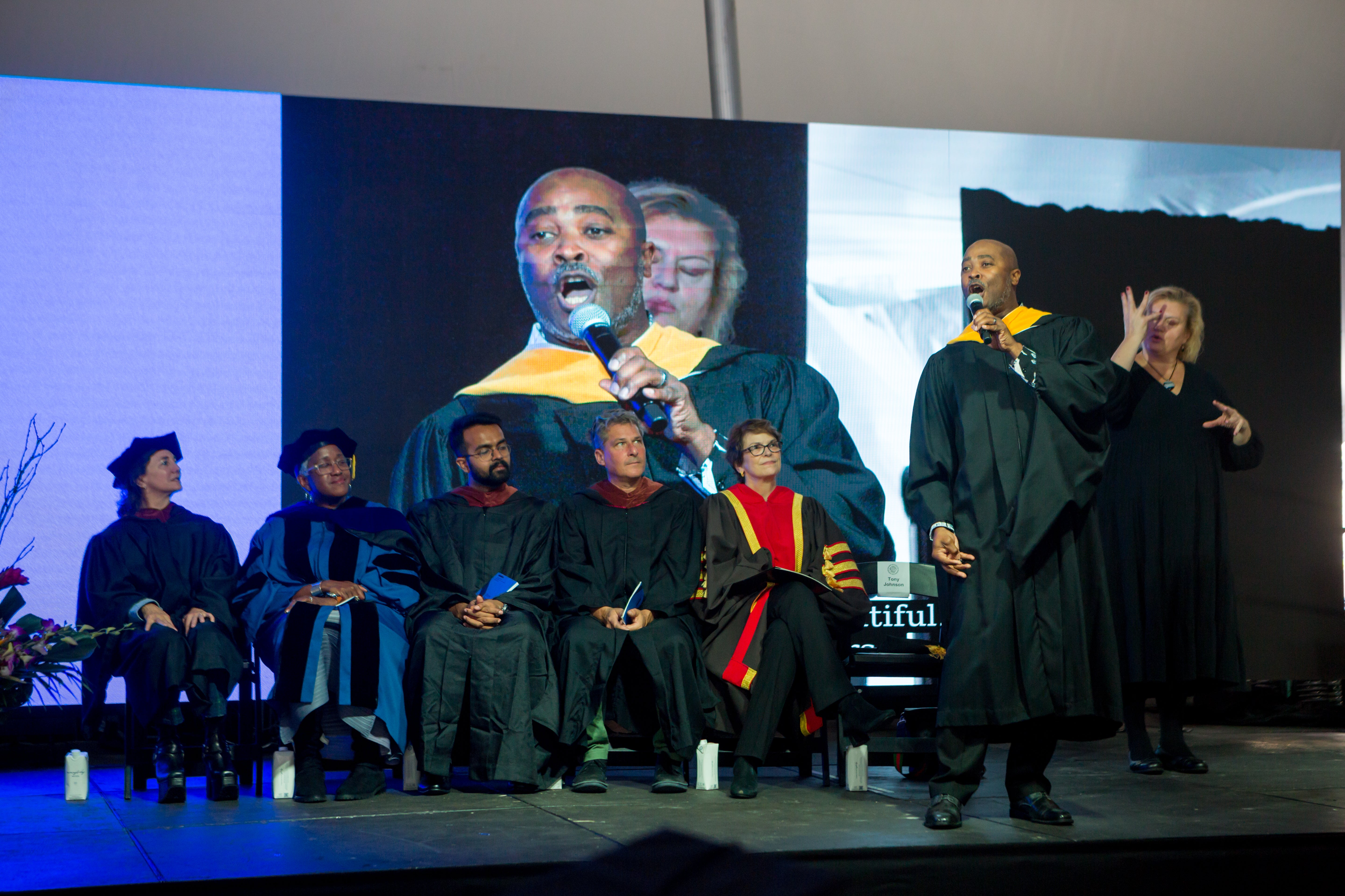 /A%20black%20man%20in%20masters%20robes%20sings%20on%20a%20stage%2C%20with%20five%20professors%20seated%2C%20looking%20on.%20A%20woman%20stands%20behind%20him%2C%20interpreting%20in%20sign%20language.