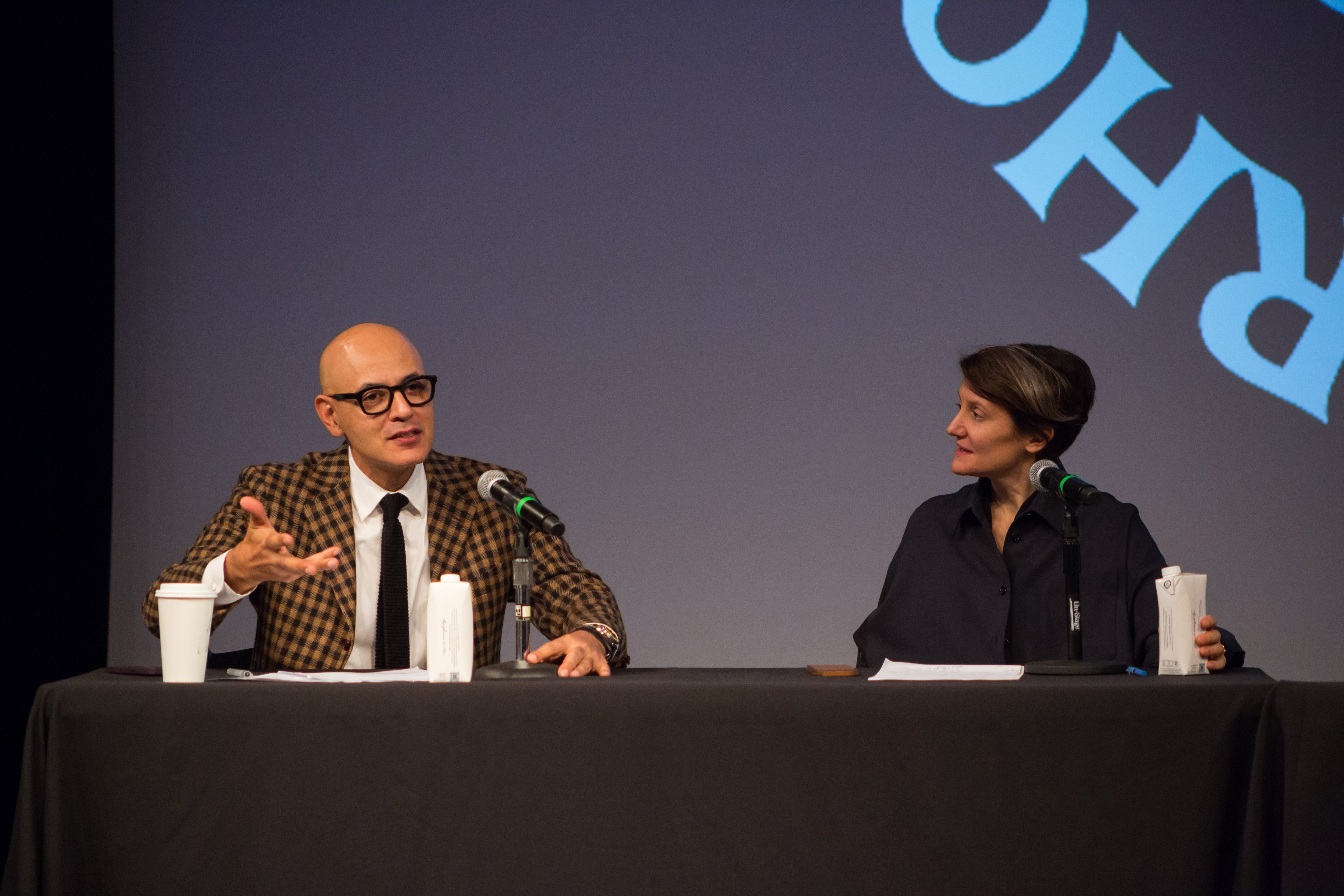 /A%20nam%20in%20a%20yellow%20and%20black%20checked%20suit%20jacket%20gestures%20and%20speaks%20into%20a%20microphone.%20A%20woman%20in%20black%20on%20the%20right%20looks%20on%2C%20listening.