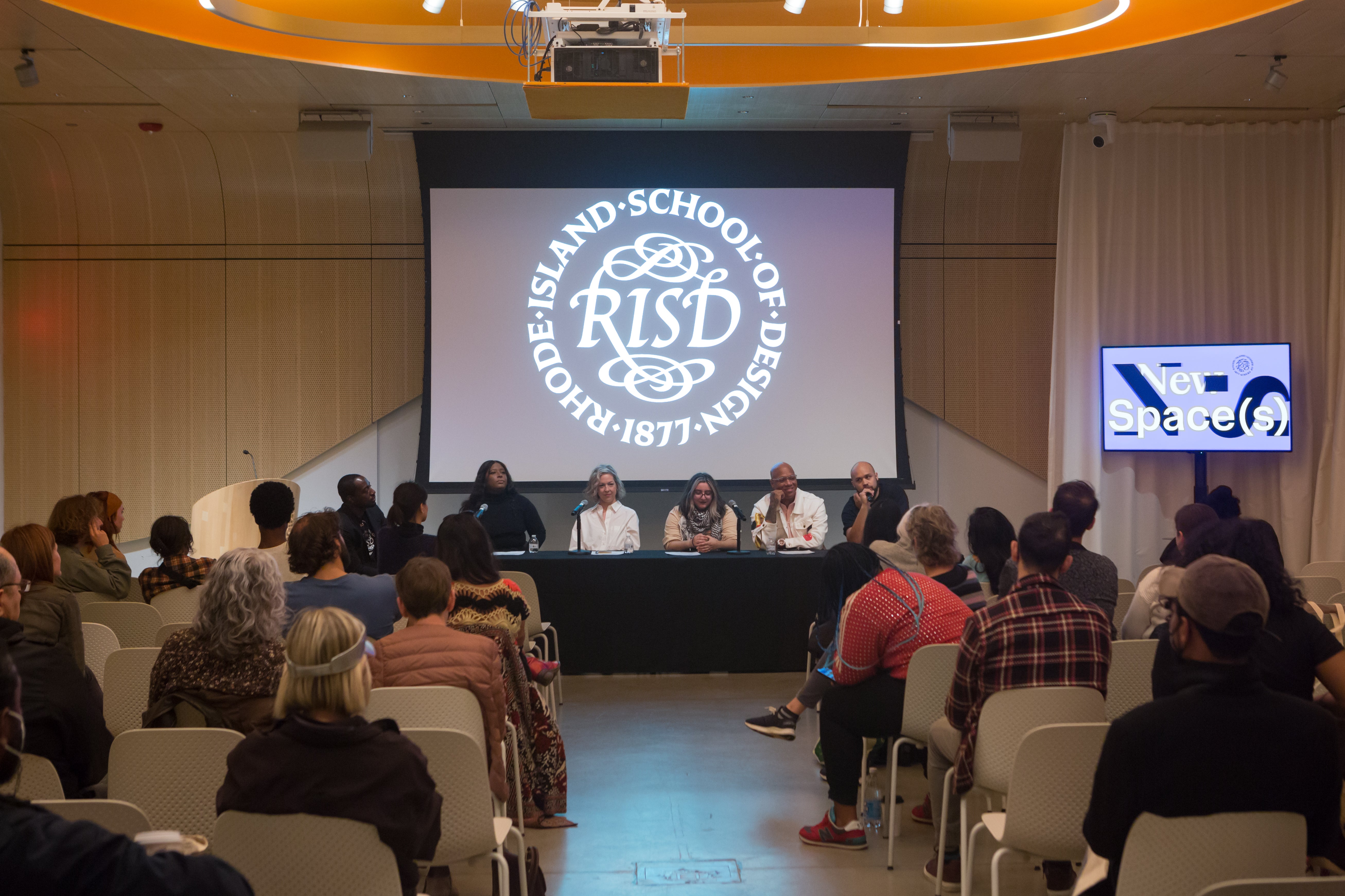 /Five%20panelists%20and%20one%20moderator%20sit%20at%20a%20table%20in%20front%20of%20a%20small%20seated%20crowd.%20The%20RISD%20seal%20is%20projected%20behind%20them.