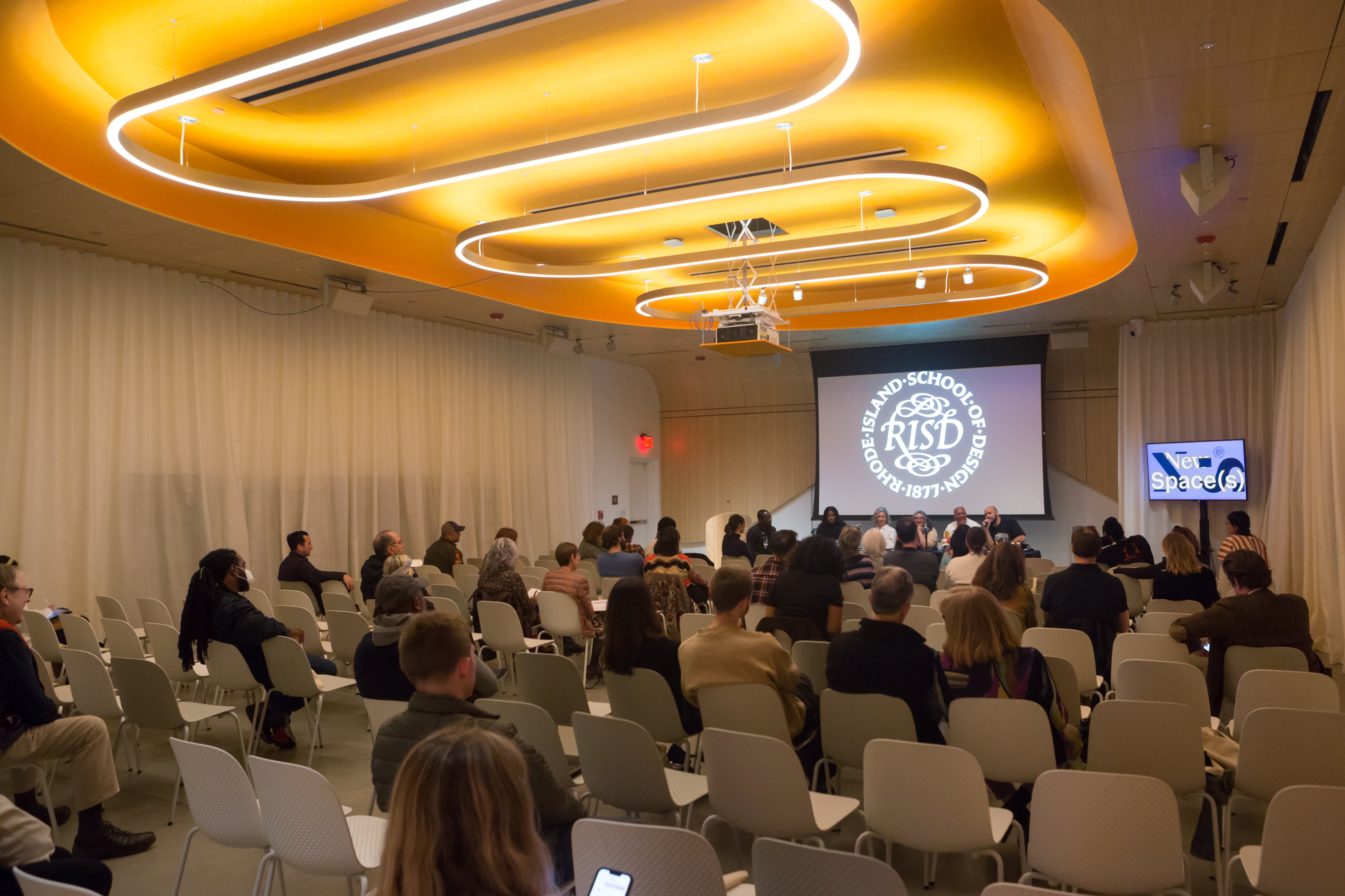 /Five%20panelists%20and%20one%20moderator%20sit%20at%20a%20table%20with%20the%20RISD%20seal%20projected%20behind%20them.%20Large%20circular%20light%20fixtures%20hang%20above%20them.
