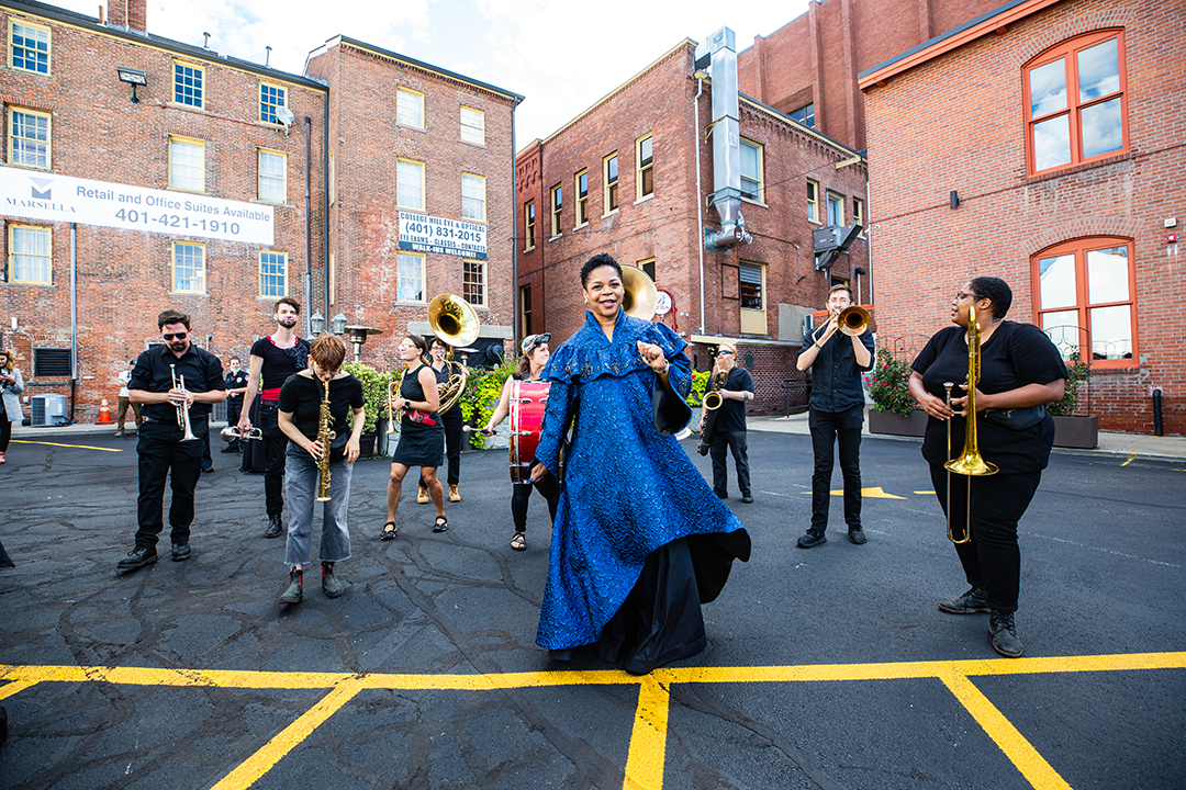 /A%20Black%20woman%20in%20blue%20ceremonial%20robes%20dances%20in%20front%20of%20a%20small%20brass%20band%2C%20performing%20in%20a%20parking%20lot%20surrounded%20by%20brick%20buildings