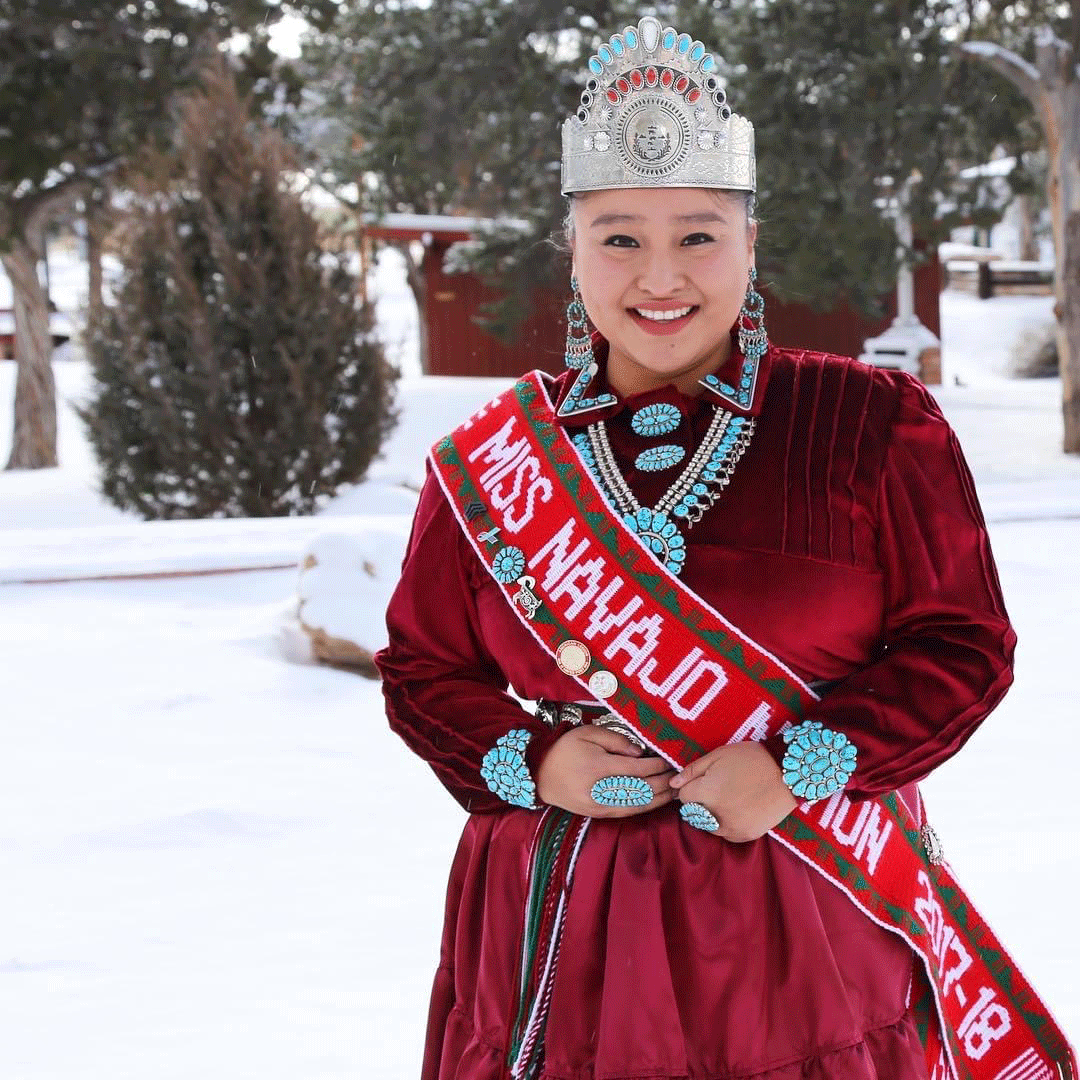 Photo of a person standing behind a snowy landscape, smiling wearing a silver crown with red and turquoise gems, and sash that reads, “Miss Navajo Nation 2017-18.” Their red velvet dress features turquoise detailing.