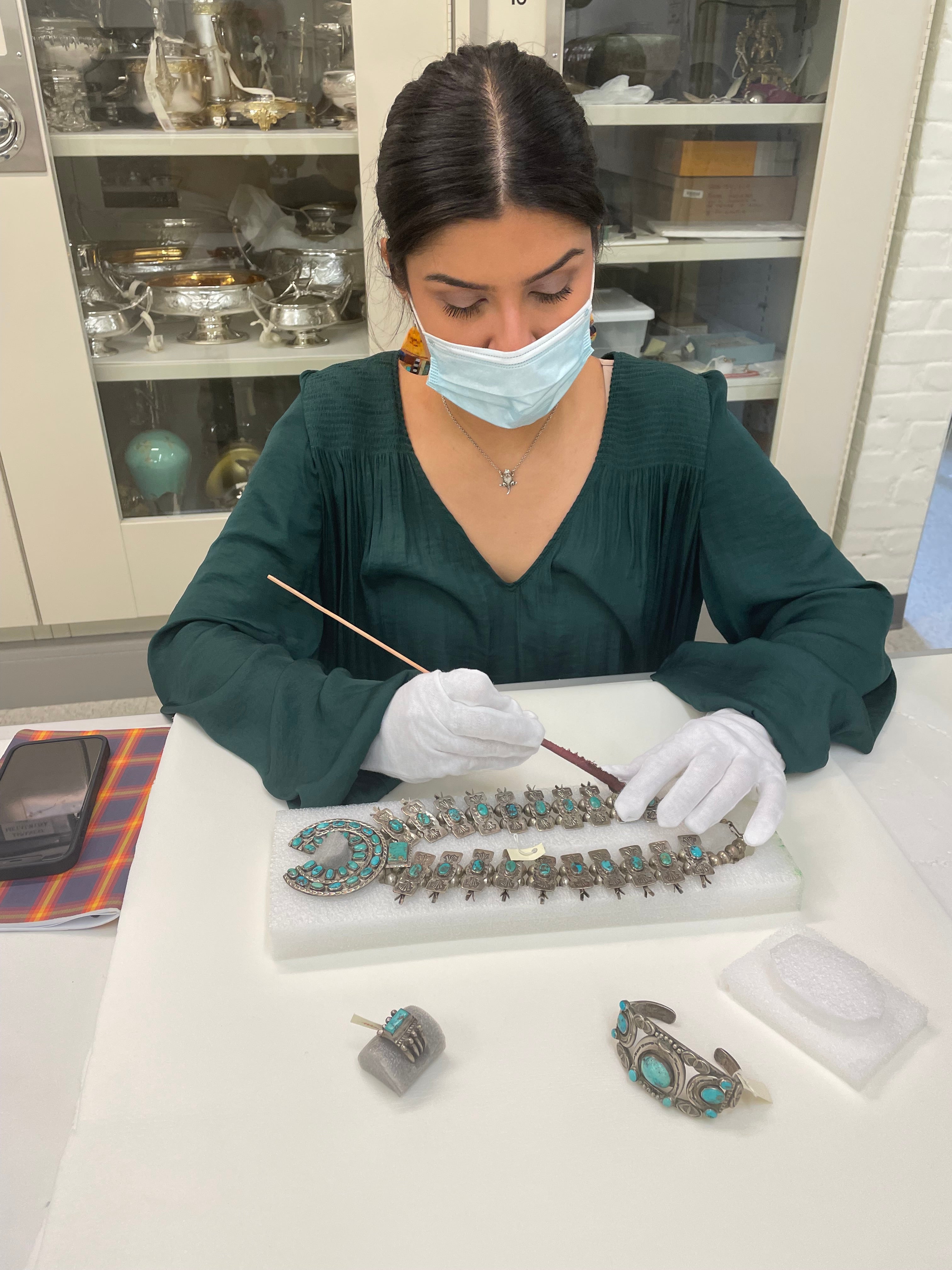 Picture of a person, wearing a mask and gloves, holding a long thin device to a silver and turquoise necklace laid in front of them on a foam rectangle.