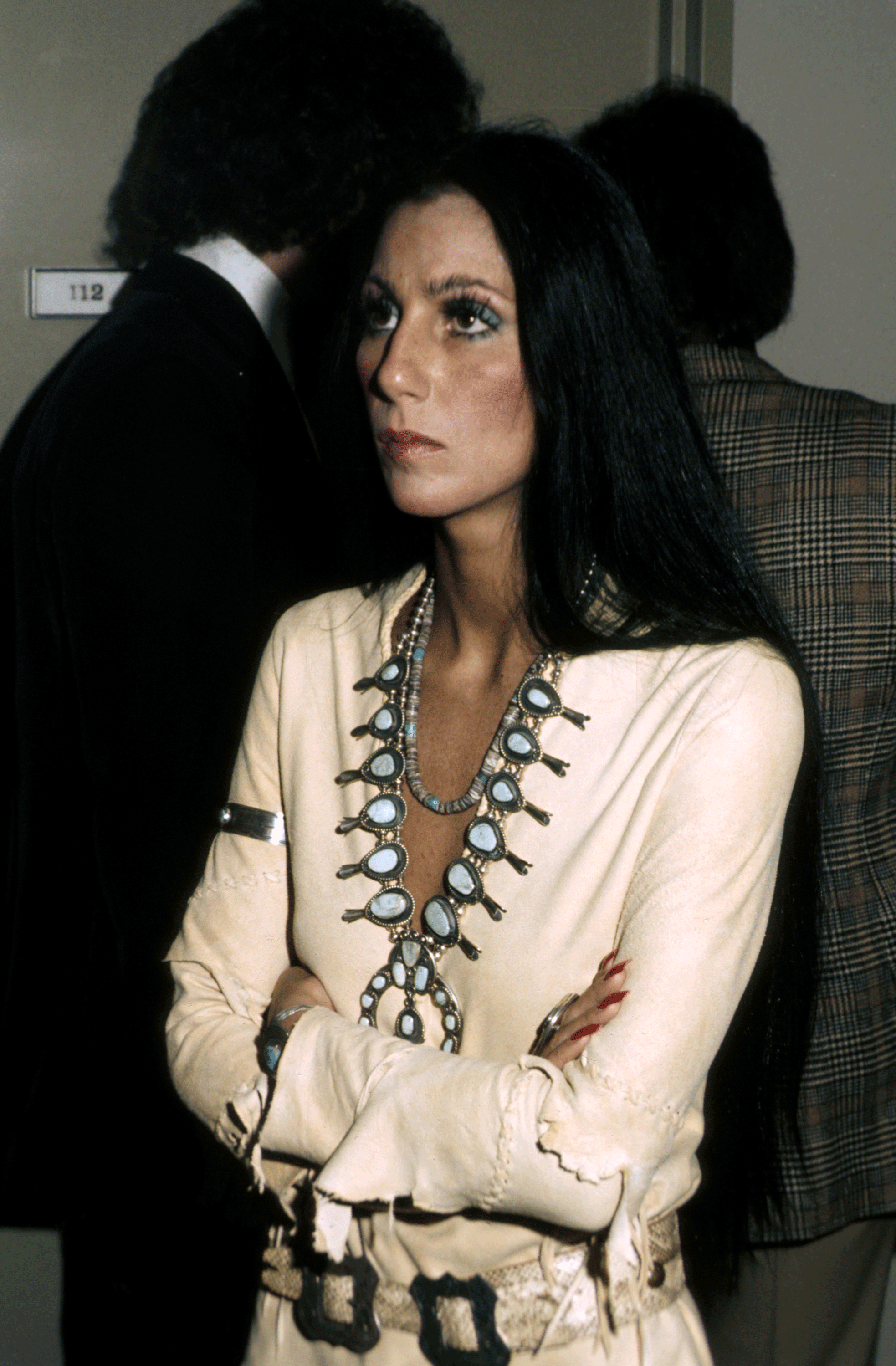 A woman with long black hair wearing white leather clothing and a silver and white pendant necklace. The person’s arms crossed, The woman is facing partly sideways with her arms crossed. 