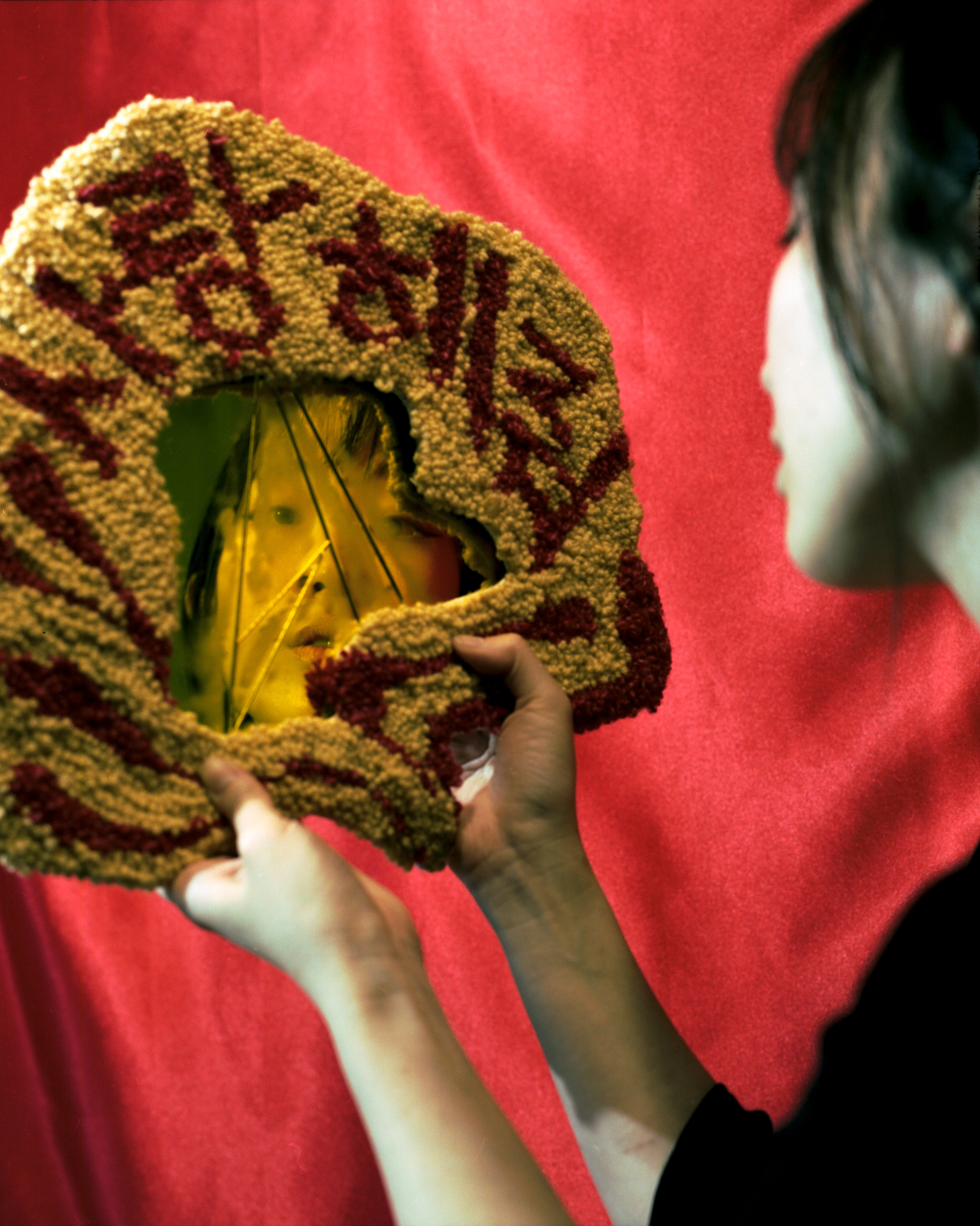 An asian woman looking into a fractured mirror covered with a tufted frame displaying the words "Love Me" in Korean.