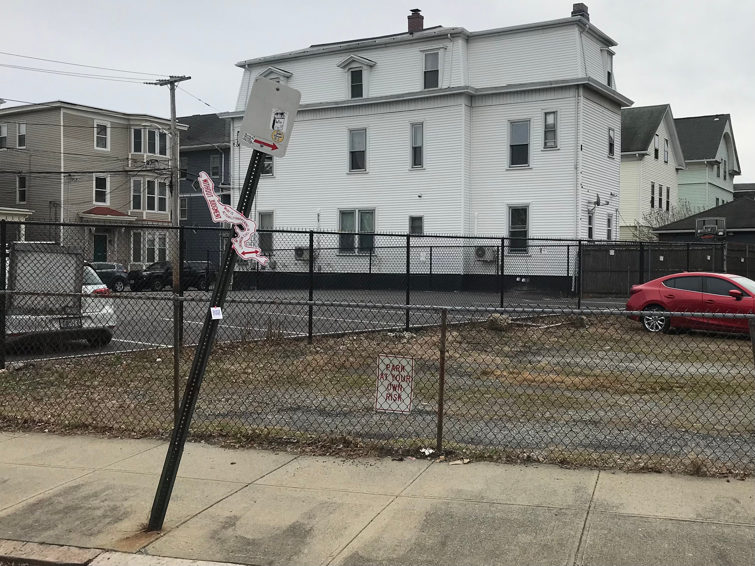 An organically-shaped red and white sign posted on a “No Parking” sign post, which reads, “Without judgment, what is actually happening in this moment?”