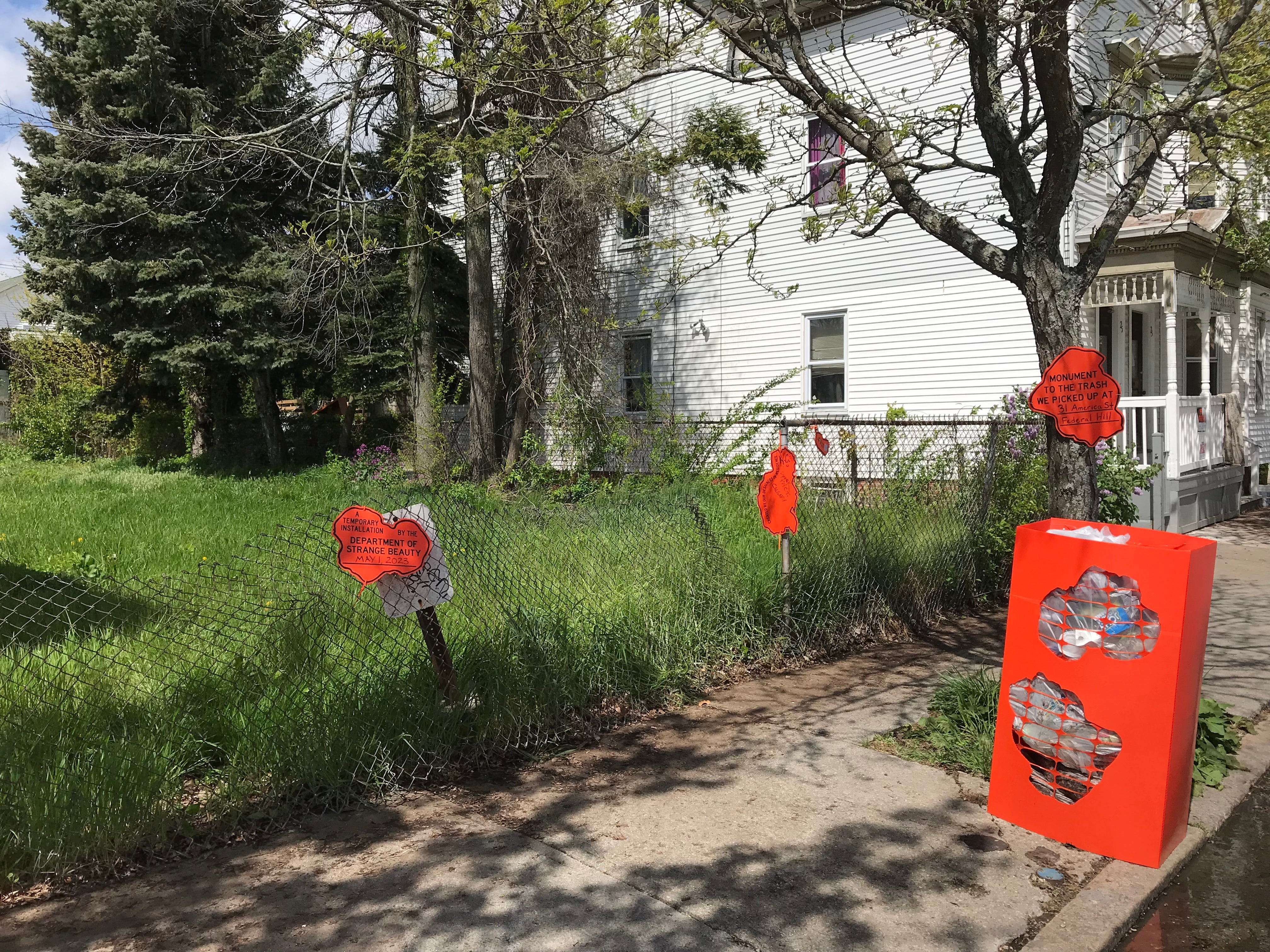 An orange box full of trash sitting on a curb next to a grassy lot, surrounded by orange signs that say, “A temporary installation by the Department of Strange Beauty May 1, 2023,” “Created by our designers in residence: K.C., Dyan K.—P.A.C.T, Calgary,” and “Monument to the trash we picked up at 31 America Street.”
