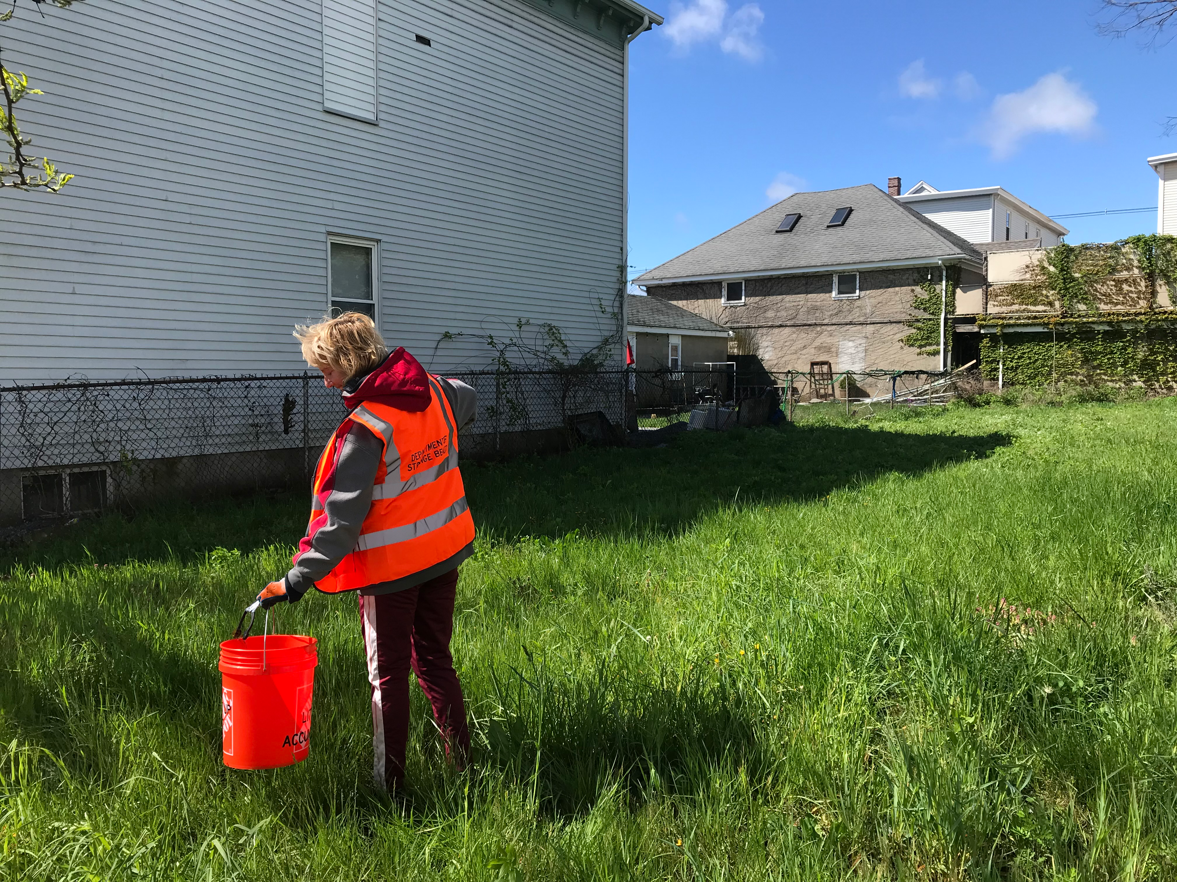 A woman in an orange vest with “Department of Strange Beauty” on the back uses a trash grabber to drop litter into an orange bucket.