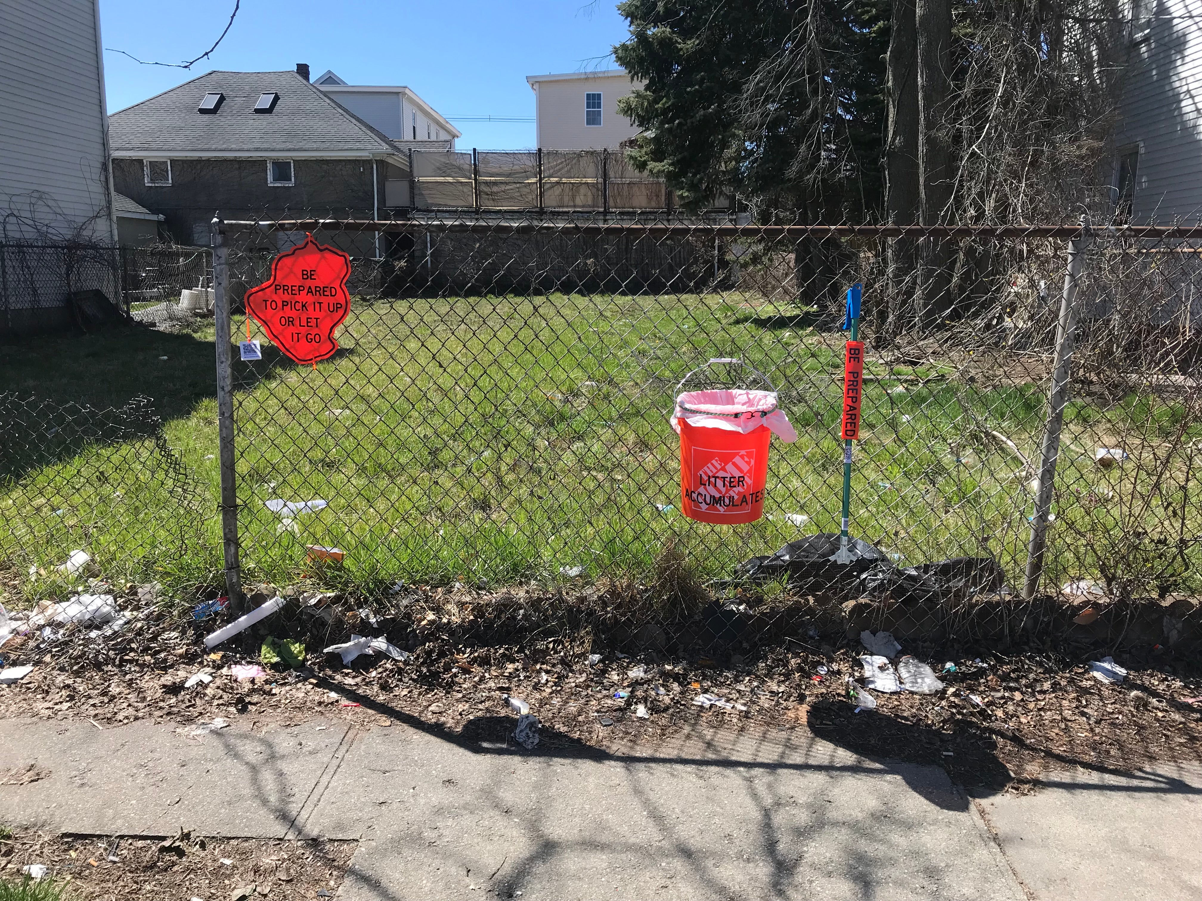 An orange sign, orange bucket, and trash grabber attached to a fence where litter has accumulated.