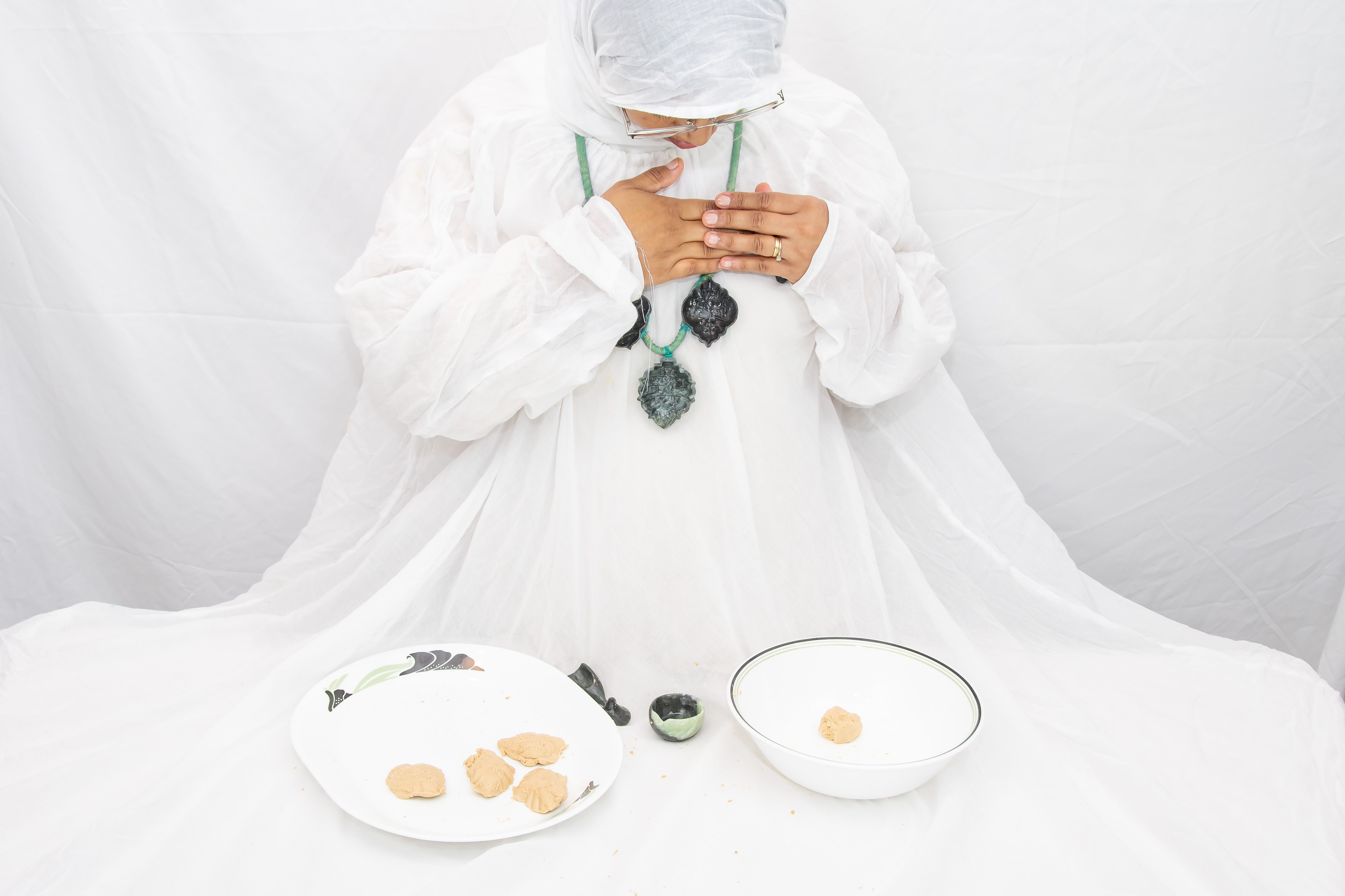 Figure with white garment draped over a table wearing a necklace with soapstone molds pressing mishti against her chest .