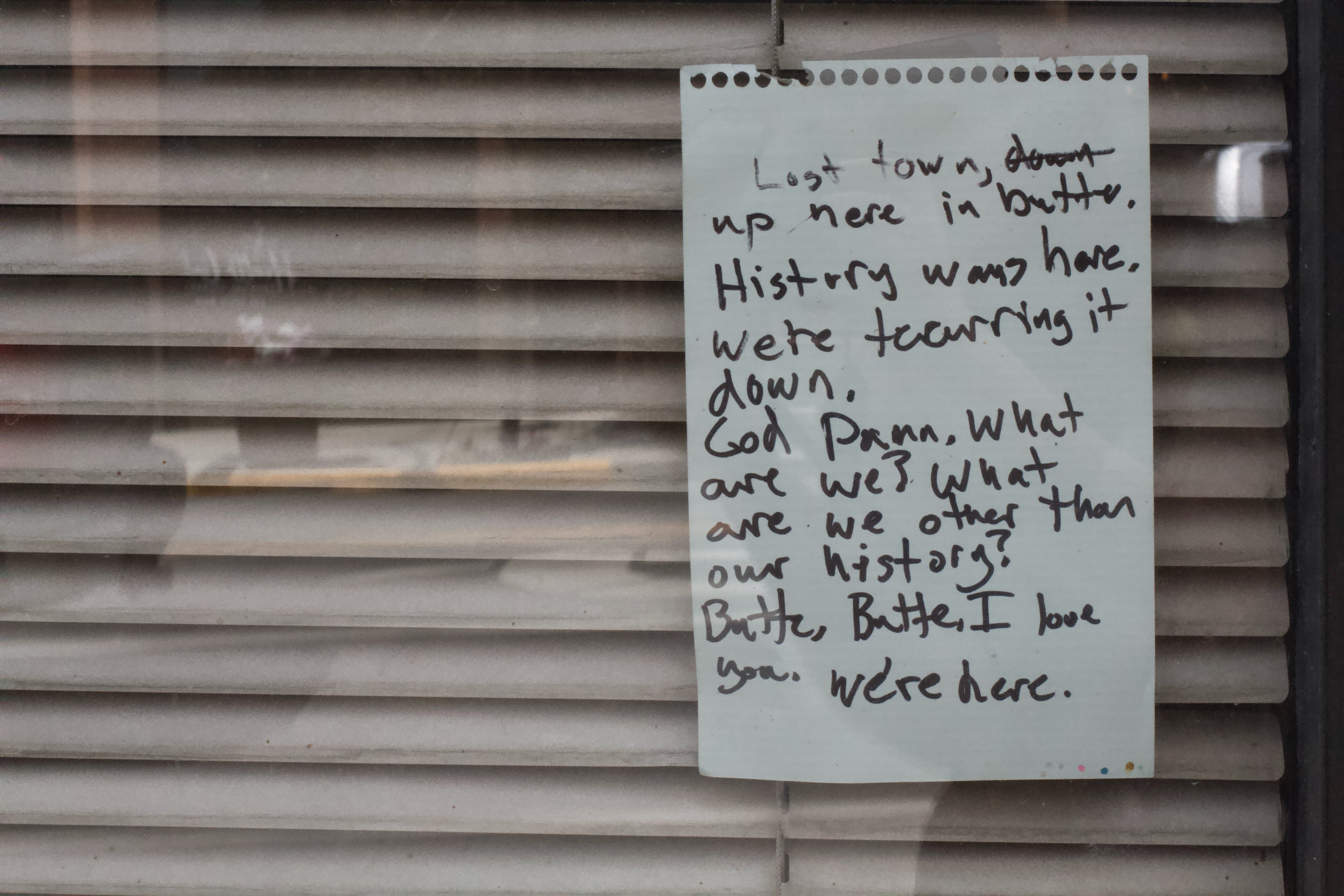 A note taped inside of a bar window that reads, "Lost town, [down is crossed out] up here in Butte. History was here. We're tearing it down. God damn, what are we? What are we other than our history? Butte, Butte, I love you. We're here." The blinds inside the bar window are shuttered.