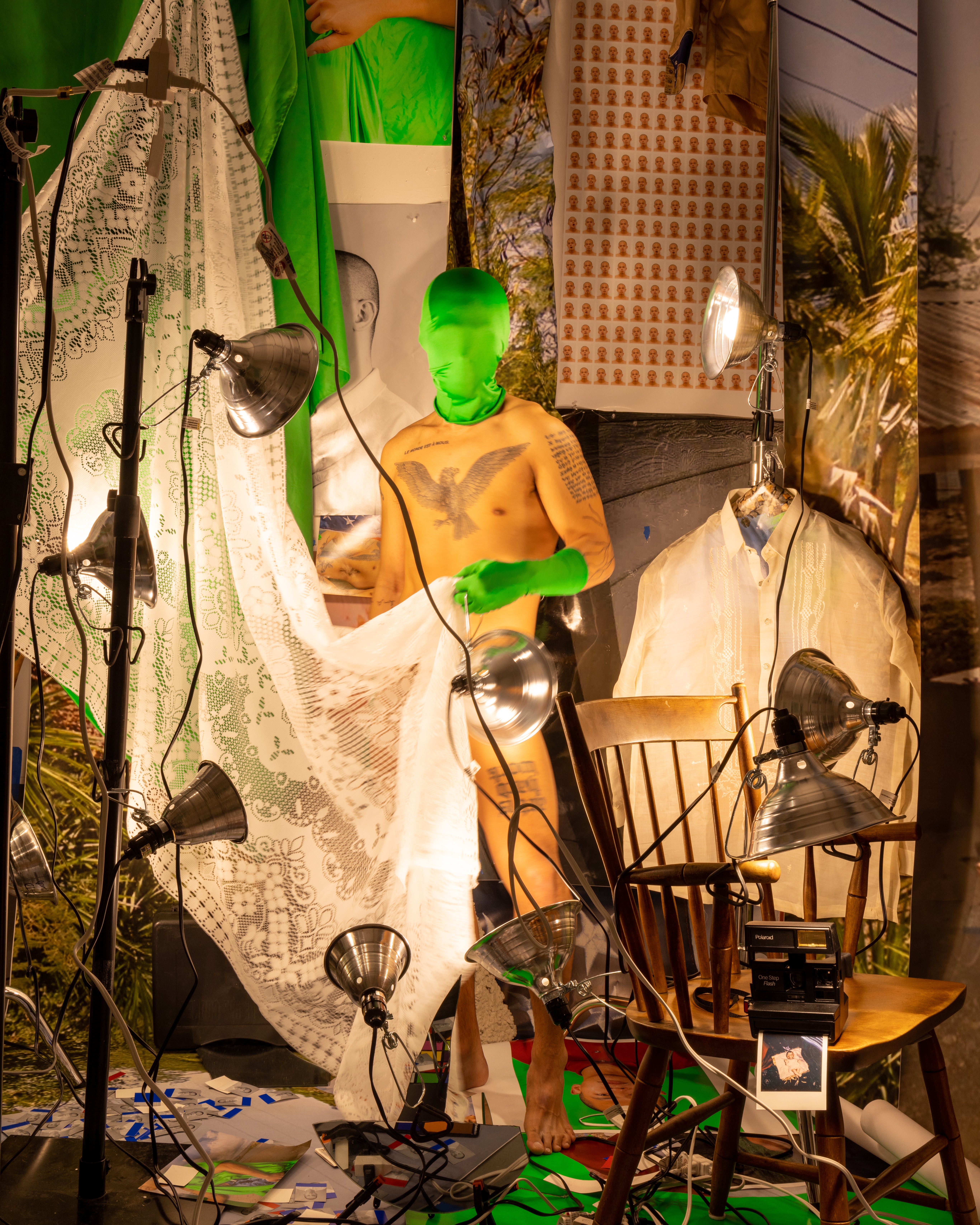 A nude man stands in a crowded room of prints and hot lights. His face is obscured by a green mask.