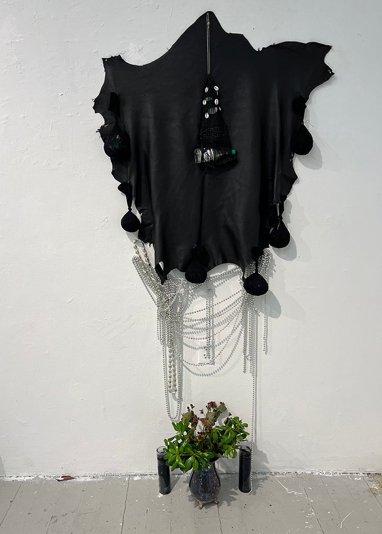 Black leather with several sacks sewn around the edges. A weaving including a glass hot sauce bottle hangs overlapping. Beneath on the wall dangles silver Mardi gras beads. Beneath on the floor are two black prayer candles, a silver vase containing spiritual herbs.