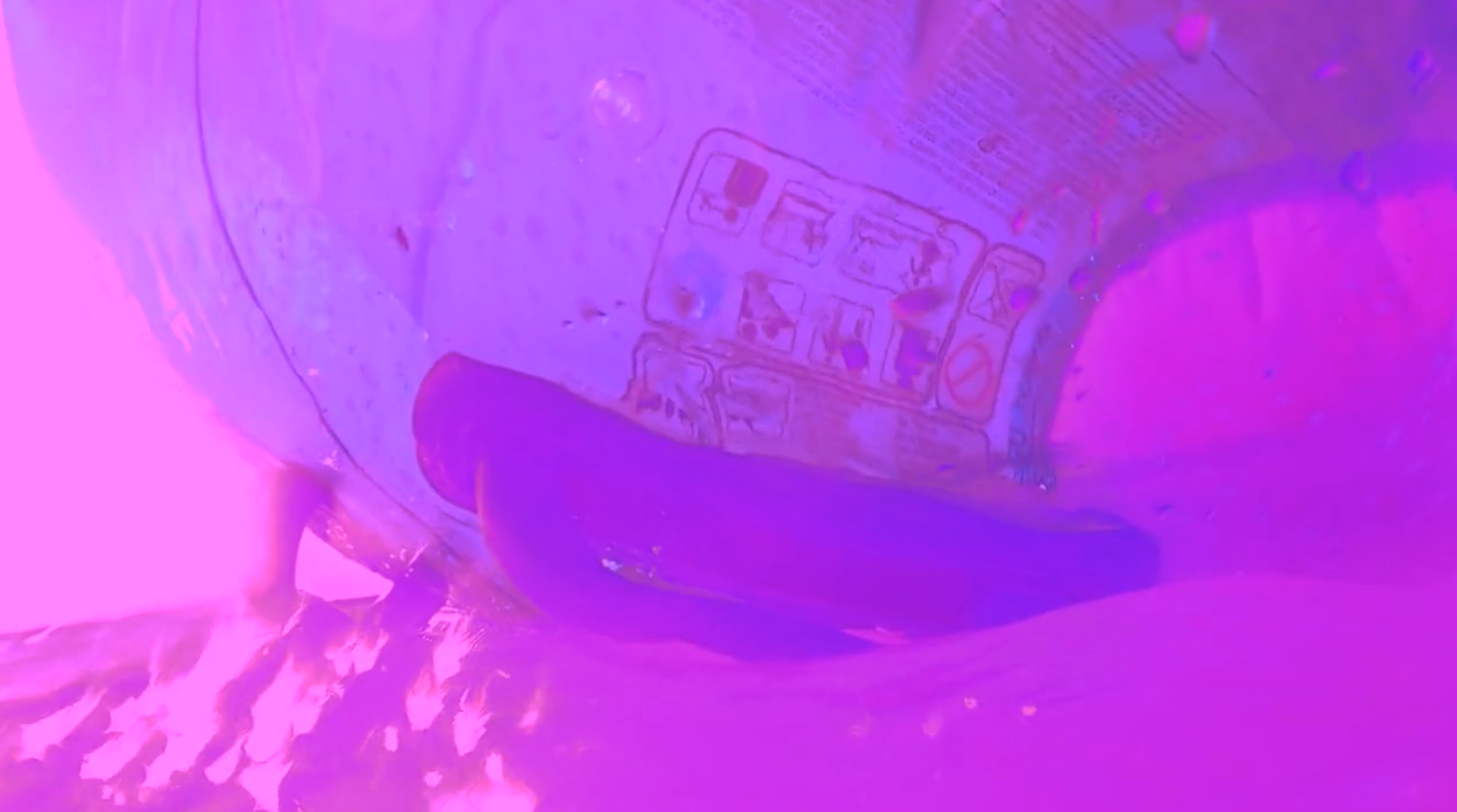 A transparent blue inflatable pool toy in the shape of a torso floats on a pink ocean. The inflatable has two handles on the back and warning text in various languages printed on its surface.