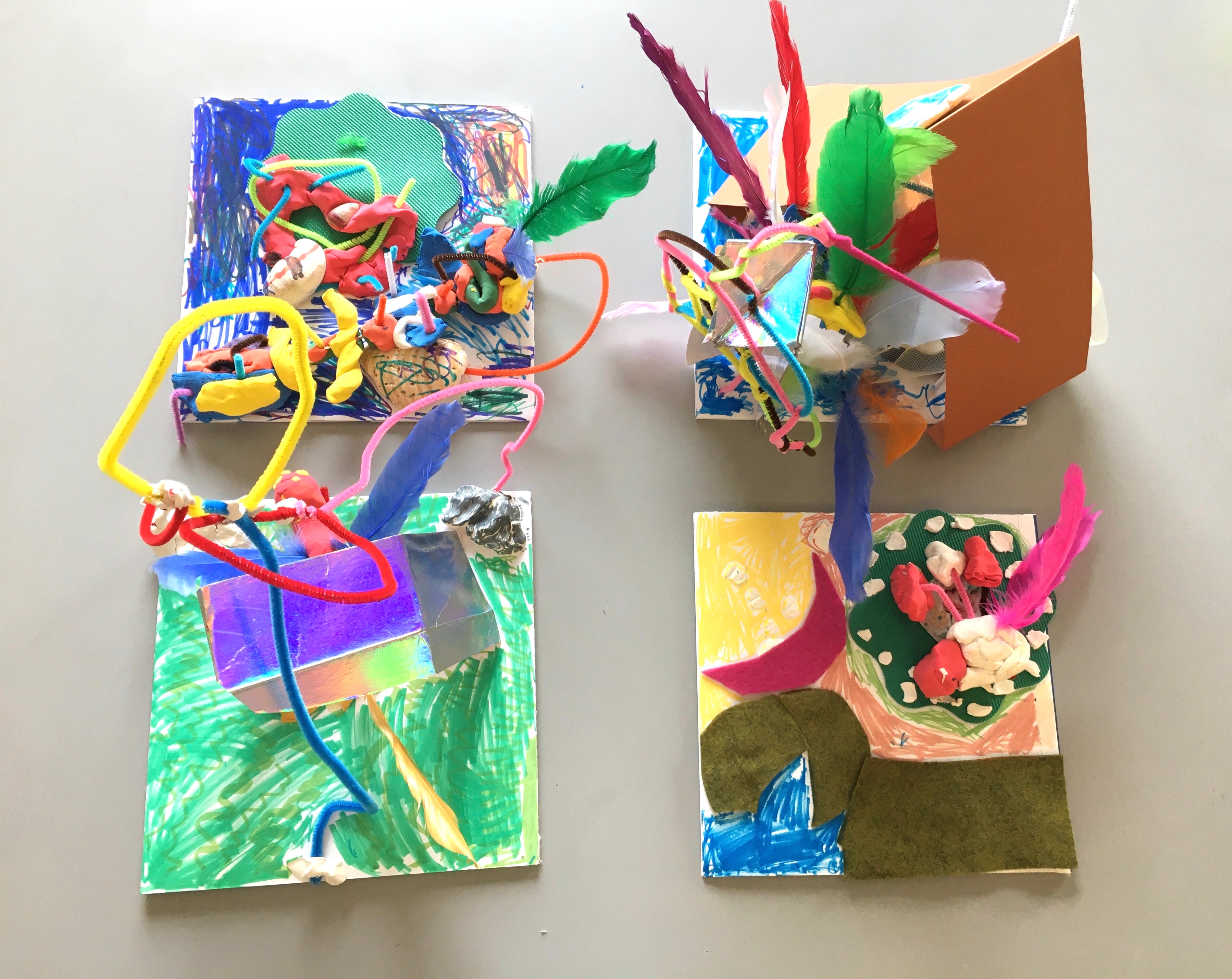 Four "habitats" built on square pieces of foam core, colorful air dry clay, pipe cleaners, shells, feathers, repurposed fabrics and paper, marker. Each habitat is constructed and colored in its own way, with some elements extending upwards towards the camera. 