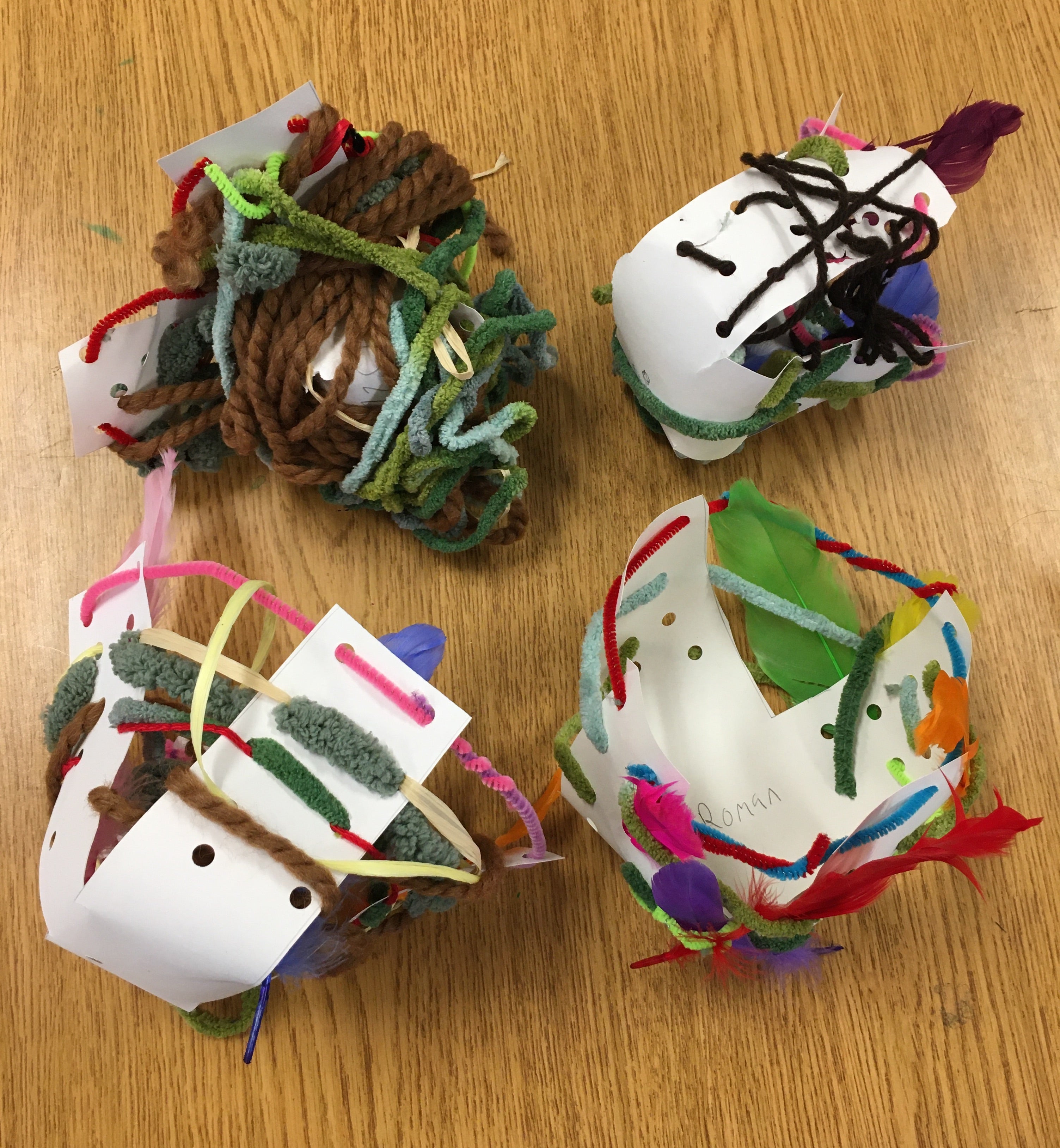Four nests made of pipe cleaners, various yarns, raffia ribbon, and feathers woven through and around cupped x-shaped frames made of hole-punched white paper. Each nest has its own style and color scheme.
