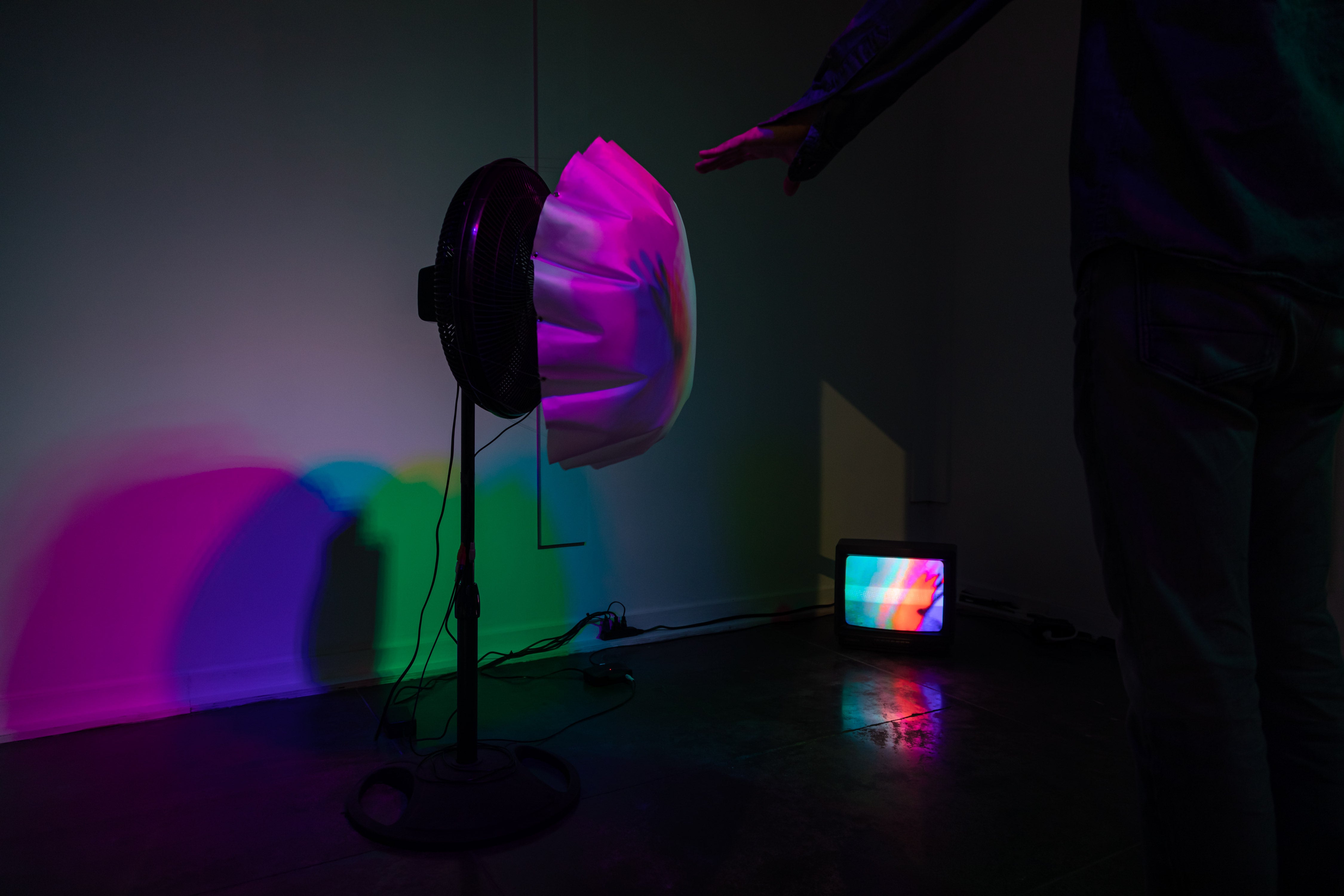 White fabric is attached to a fan and being blown up under colorful light.