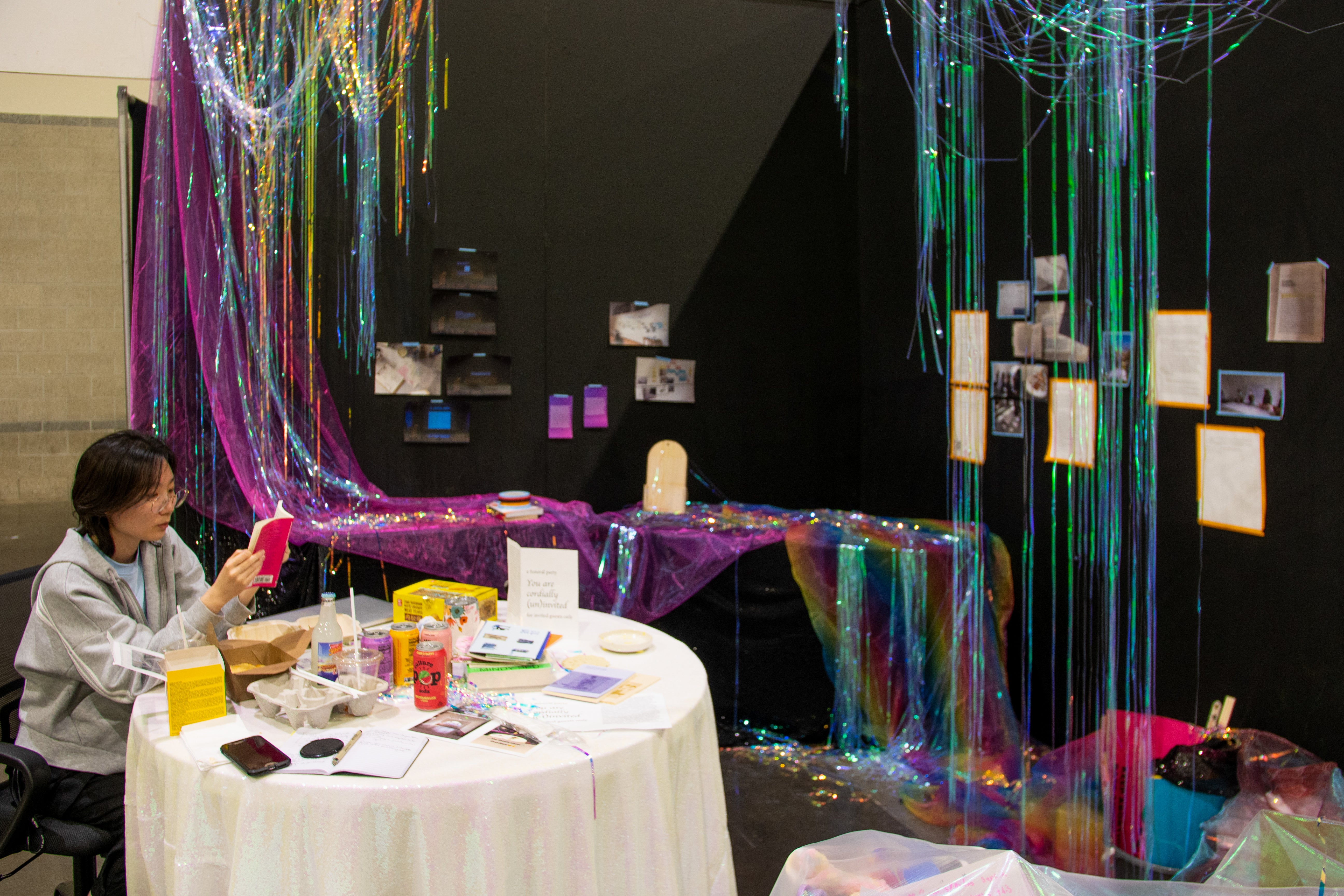 books, food, and drinks on white table; iridescent party streamers hanging from above like curtain; photos and writings on corner-shaped black walls