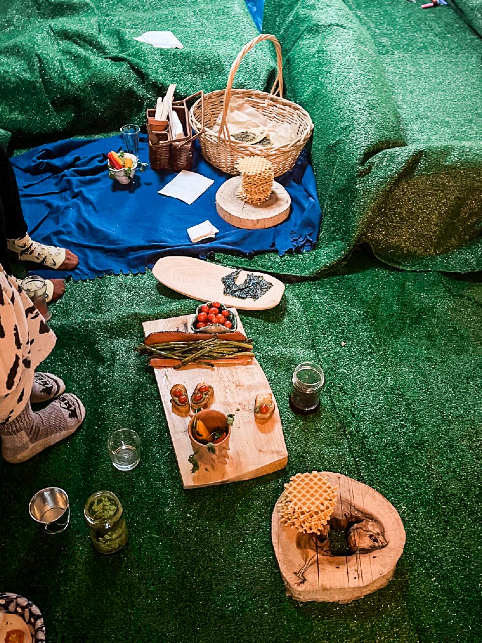 The green astroturf-covered floor of the apartment used for the indoor picnic. A blue blanket with a basket and a stack of waffle cookies, and other wooden food platters.