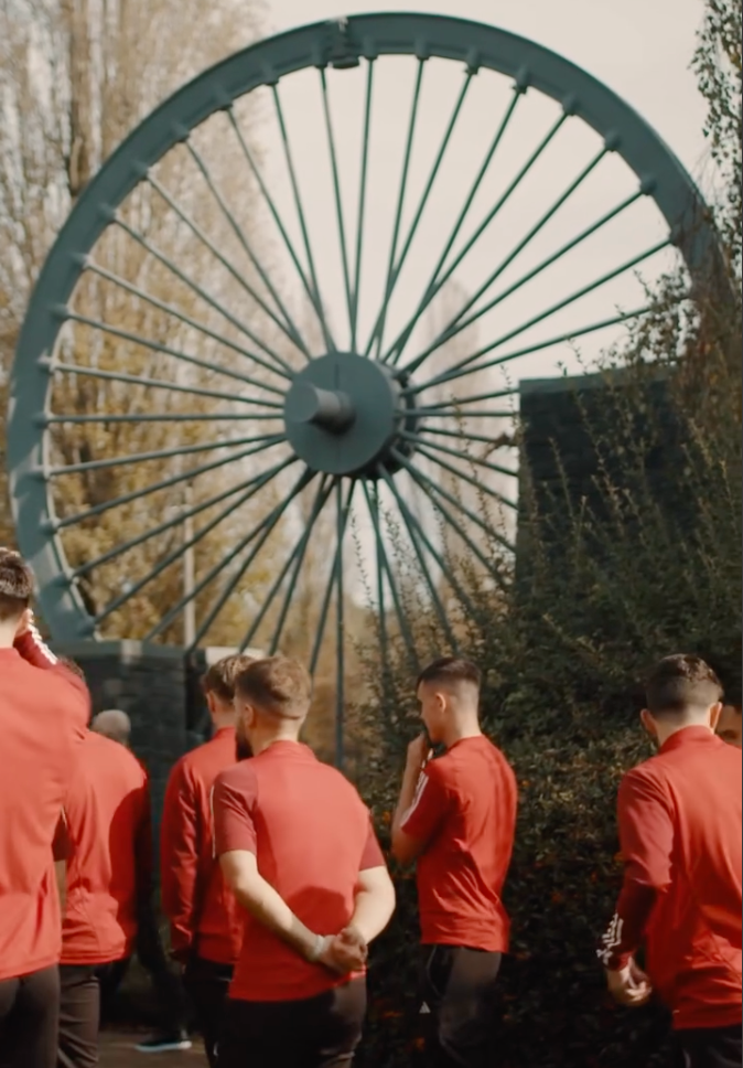 Welsh football players standing in front of a colliery wheel memorial to the Gresford mining disaster.