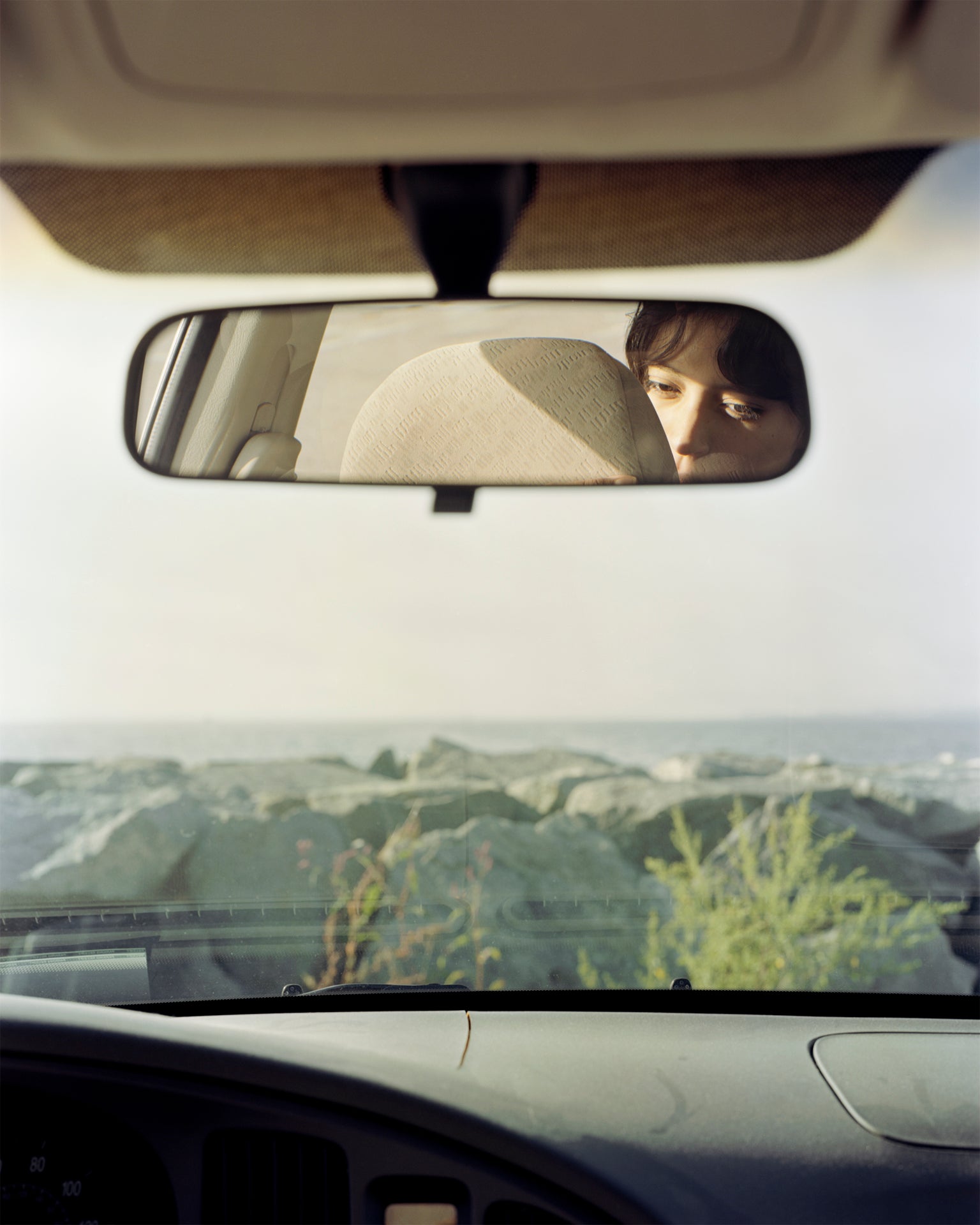 /Photograph%20from%20inside%20of%20car%20looking%20out%20through%20the%20windshield%20to%20a%20rocky%20beach.%20The%20dashboard%20sits%20at%20the%20bottom%20of%20the%20image.%20There%20is%20a%20rearview%20mirror%20with%20a%20reflection%20of%20person%20face.