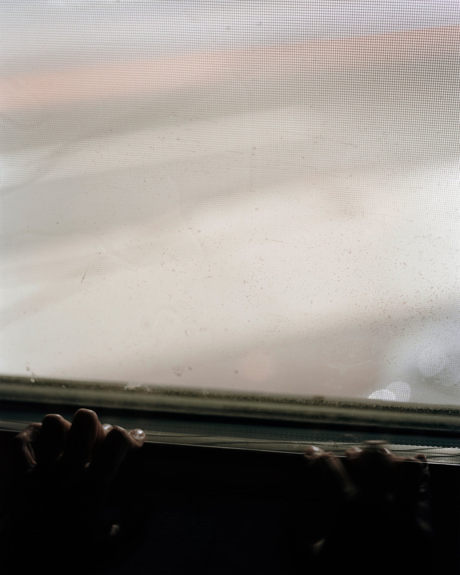 /A%20long%20exposer%20photograph%20of%20a%20hands%20resting%20a%20on%20a%20white%20windowsill%20at%20night.%20The%20hands%20are%20mostly%20in%20shadow.%20Through%20the%20window%20is%20the%20gray%20street%20with%20long%20obscured%20car%20lights.%20