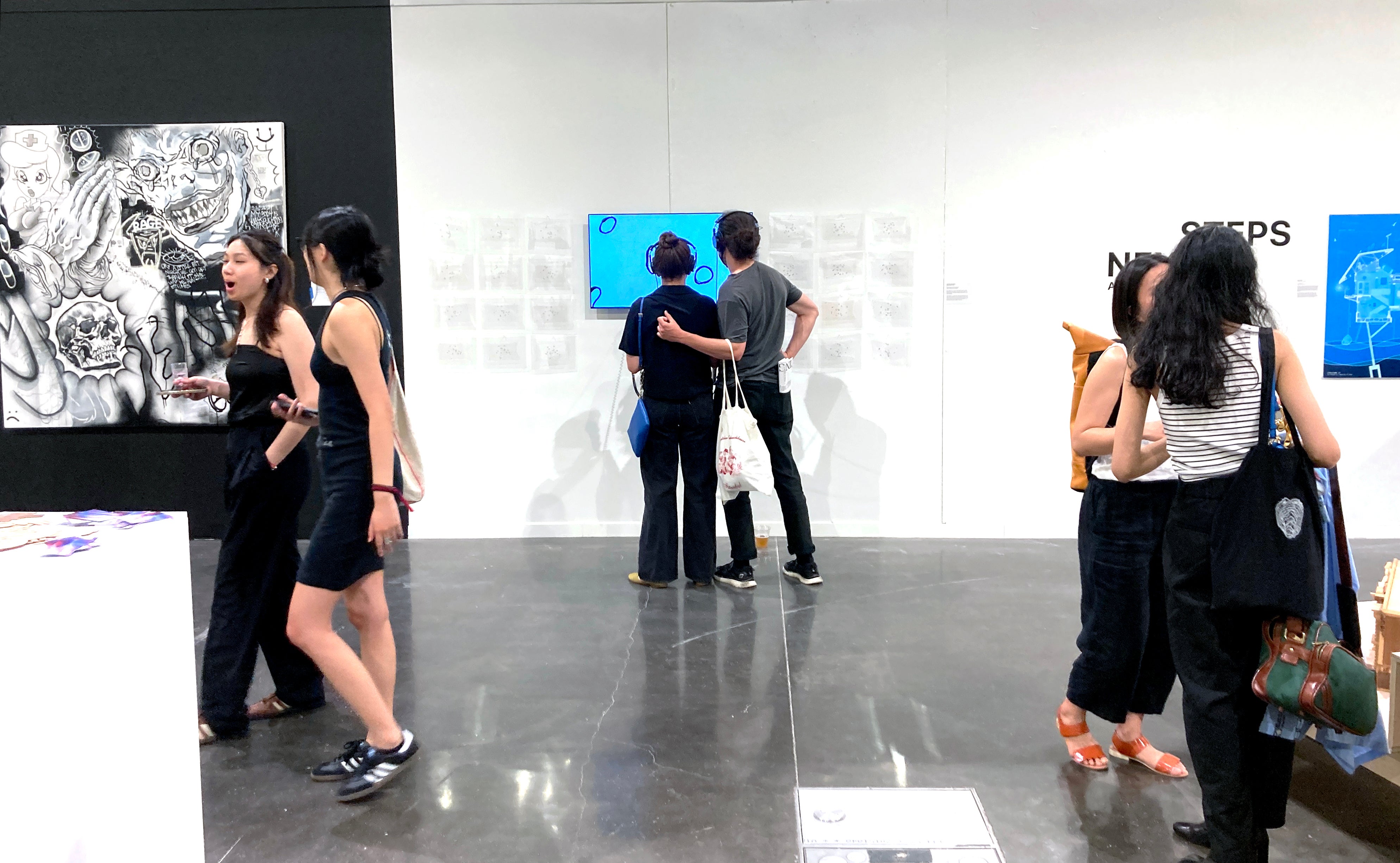 A couple, both wearing headphones, watch a film playing on screen with shapes moving against a blue sky. The man has his arm around the woman. People bustle around them.