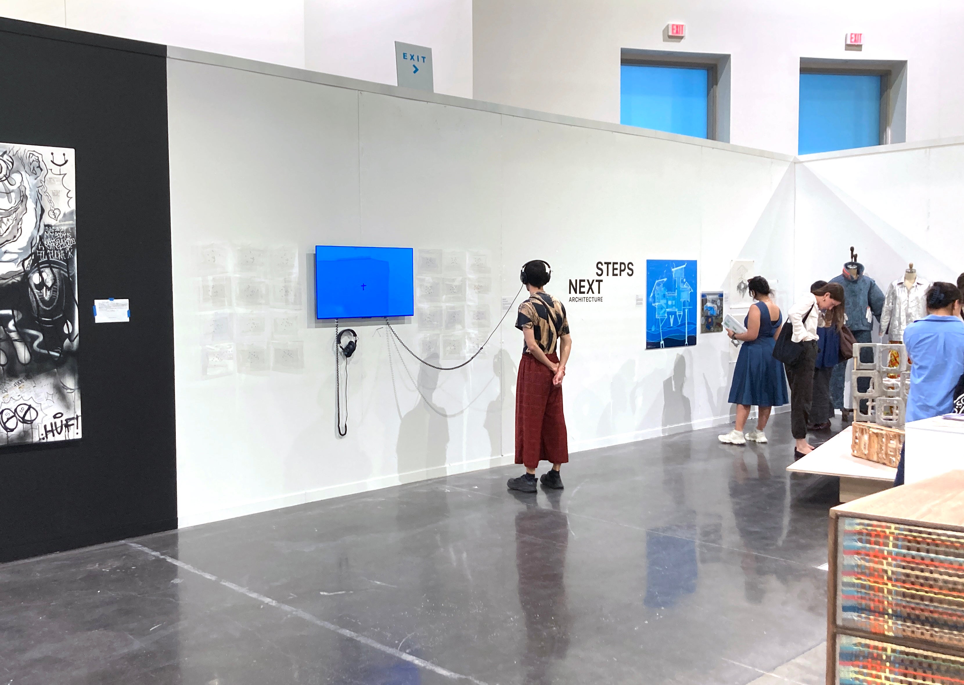 A single man reads the text on the wall next to a film installation. He is wearing headphones attached to the installation. People look at other art in the background.