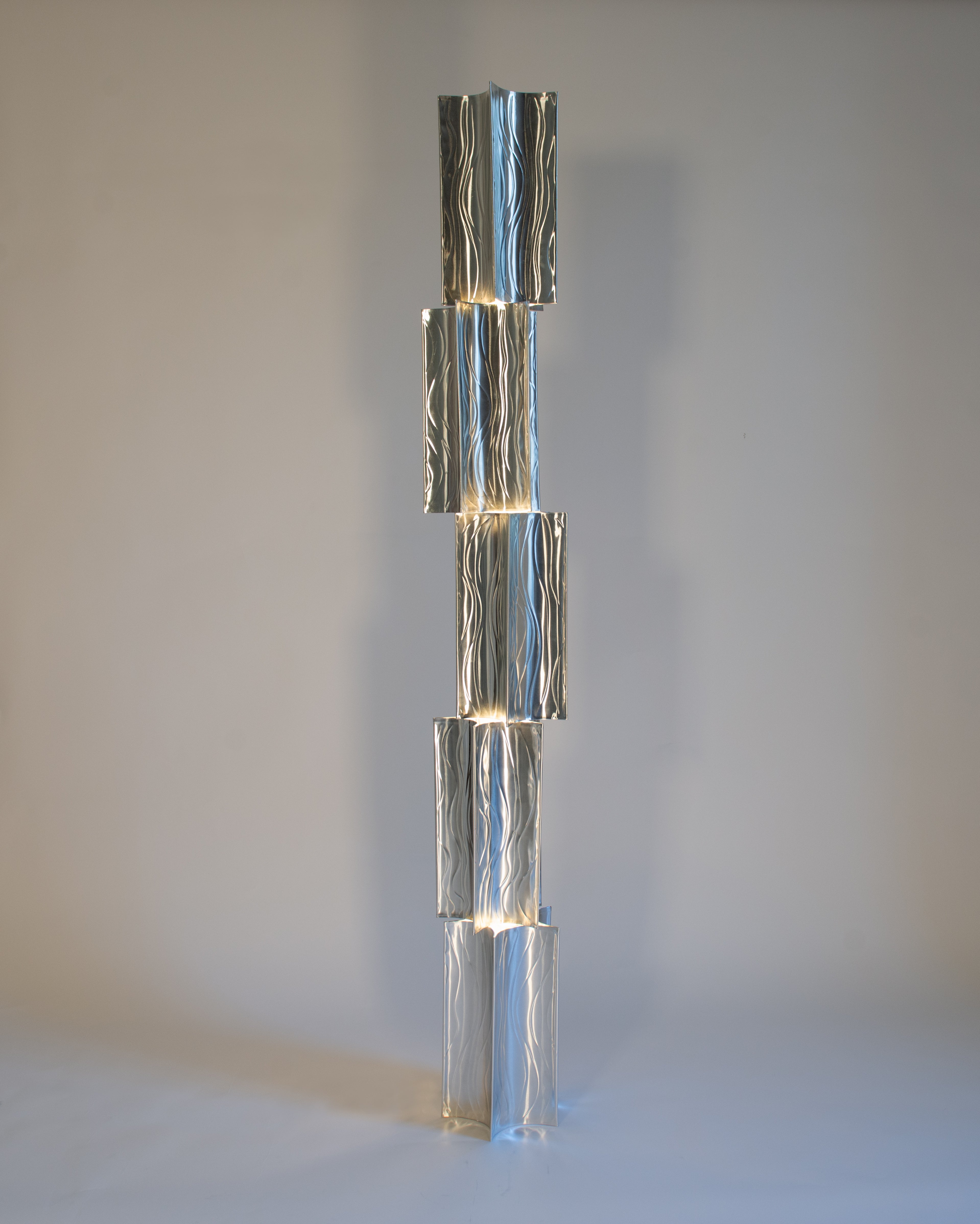 A sleek, modern floor lamp composed of a vertical stack of five aluminum lanterns. Each panel has a wavy texture and emits a subtle, warm light from the edges, creating a cascading effect.