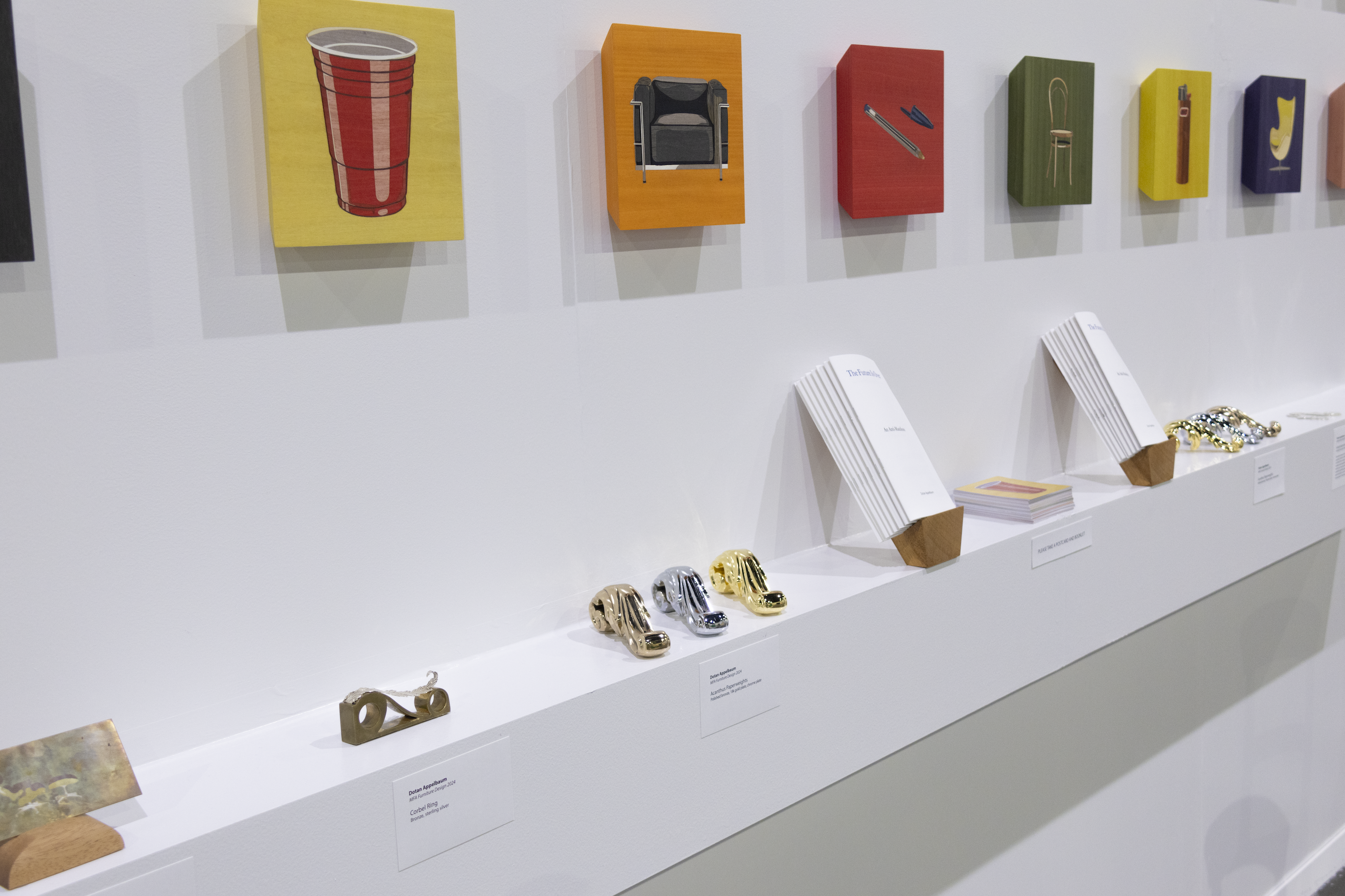 Left-side view of a shelf with Acanthus Paperweights, Corbel Ring, a decorative card scraper, booklets, and a stack of postcards. Above the shelf are colorful panels depicting chairs and ubiquitous products. 