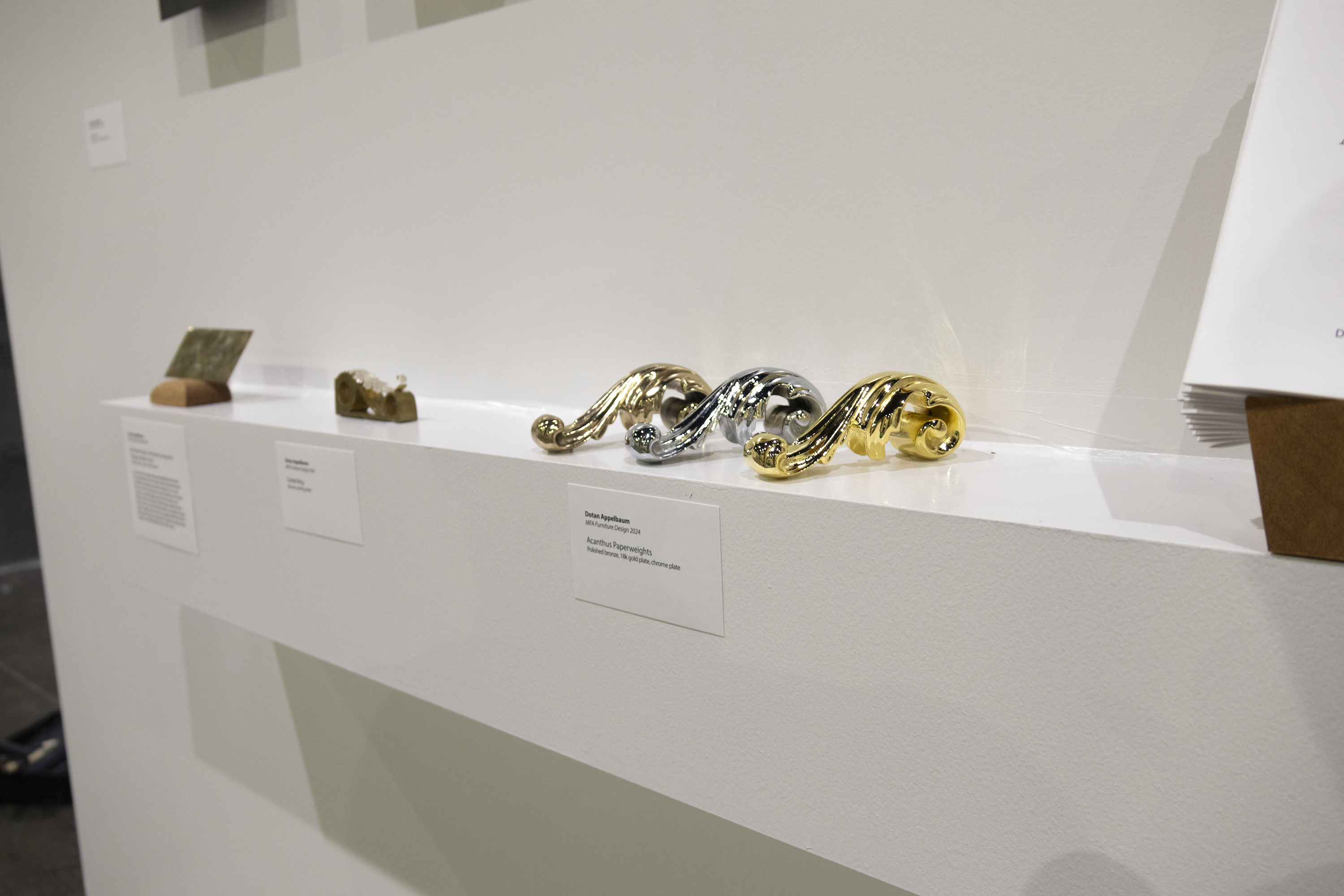 Right-side view of a white wall-hung shelf with Acanthus Paperweights, Corbel Ring, and a decorative card scraper. Title cards are posted on the front face of the shelf.