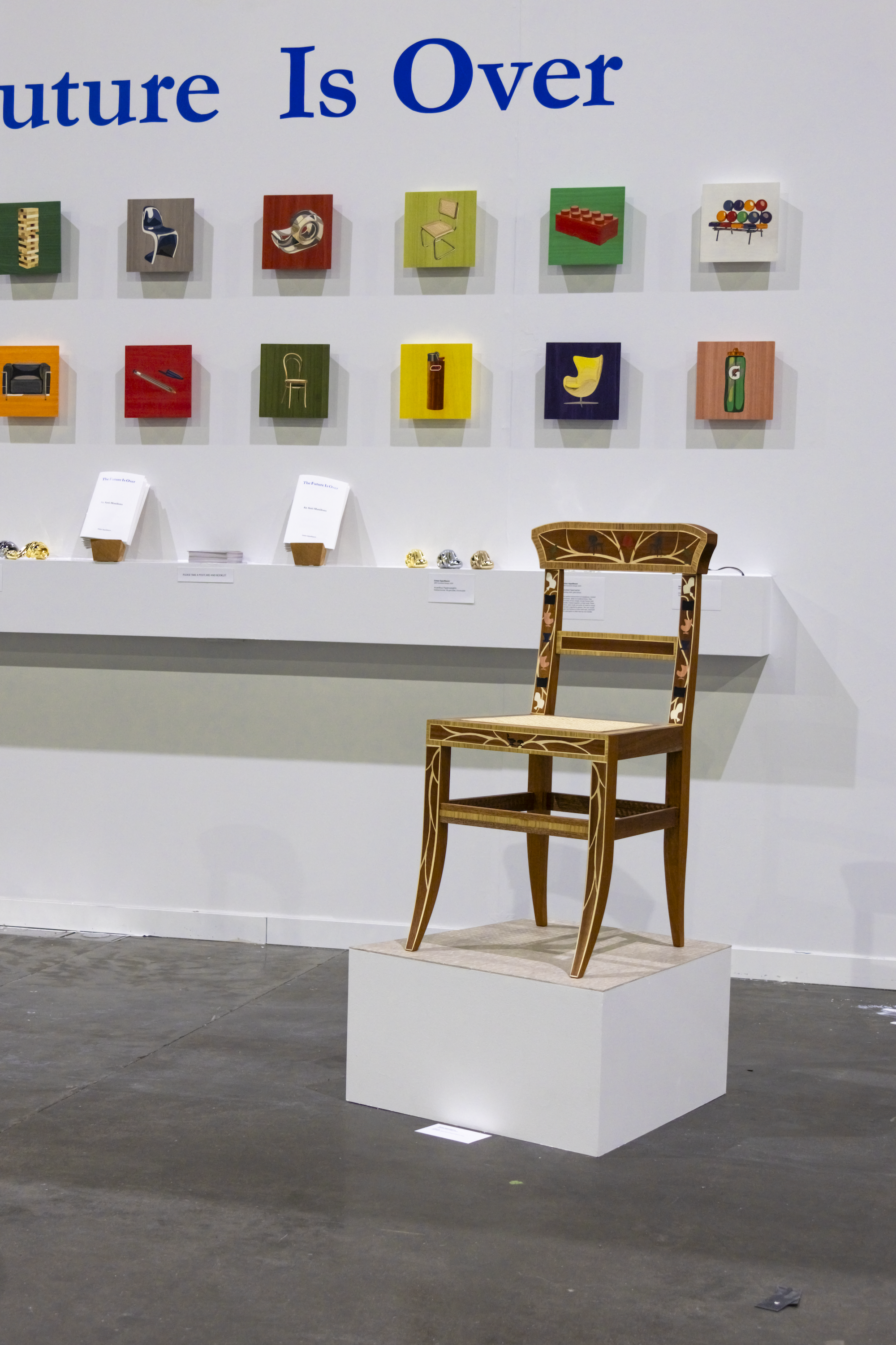 A chair with colorful inlaid depictions of chairs sits on a trapezoid-shaped pedestal with a linoleum top surface. Behind the chair is a wall with a shelf, colorful panels, and blue text.