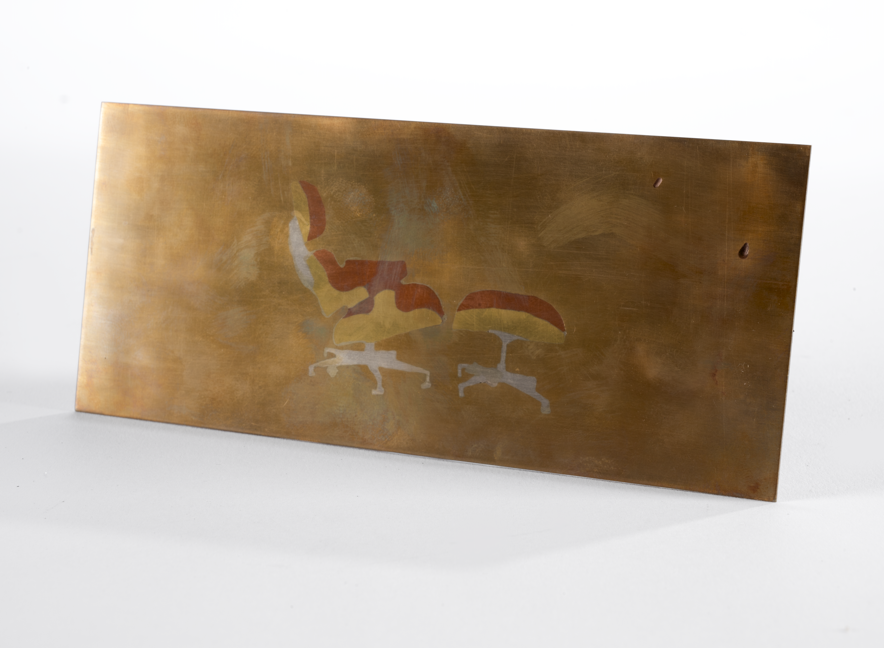 A thin rectangular sheet of bronze with an image in the center made of copper, brass, and silver depicting the side profile of the Eames Lounge Chair and Ottoman.
