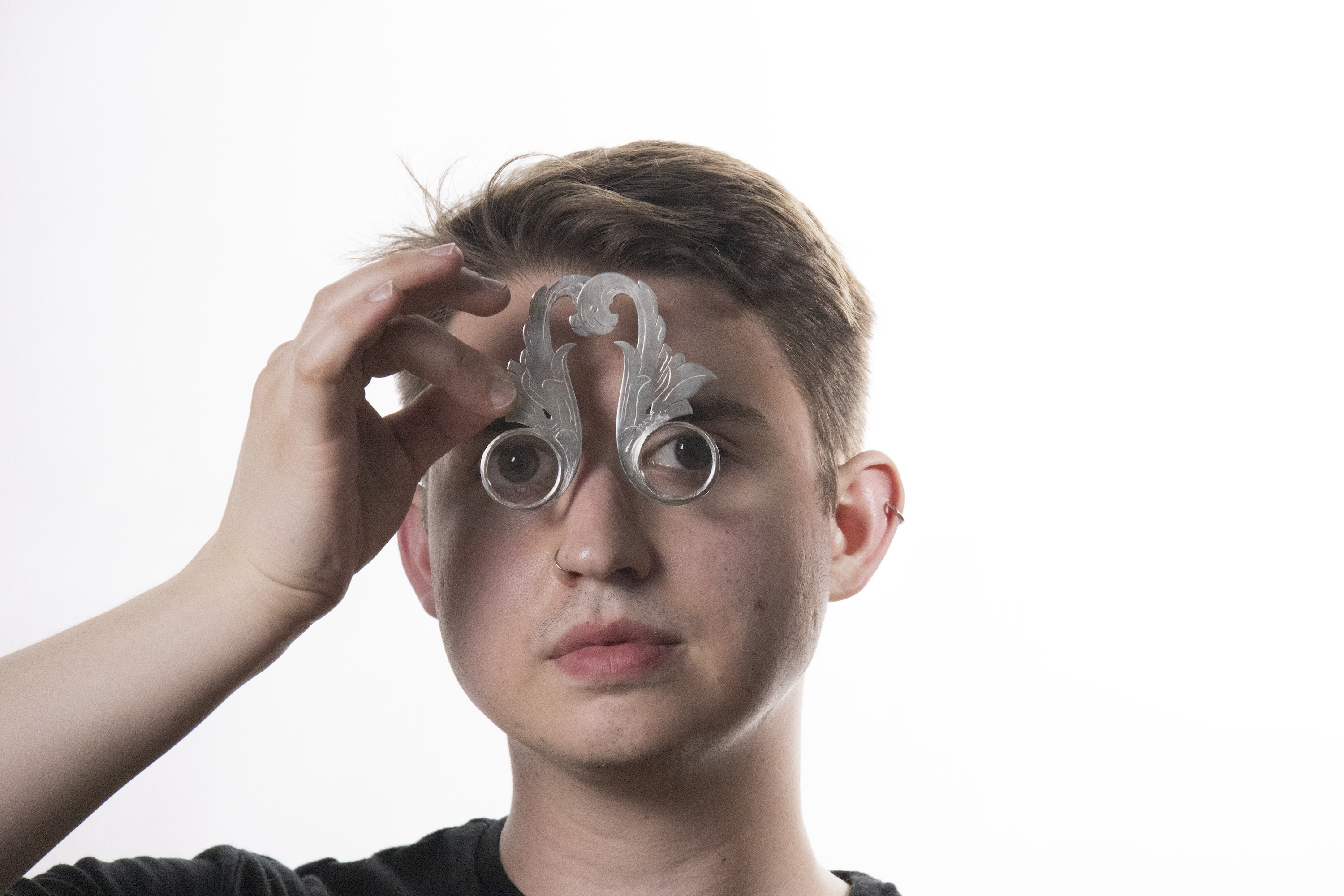 A man against a white background holds two magnifying glasses to his eyes held in silver frames that are cut and engraved to look like acanthus leaf illustrations and rivited together