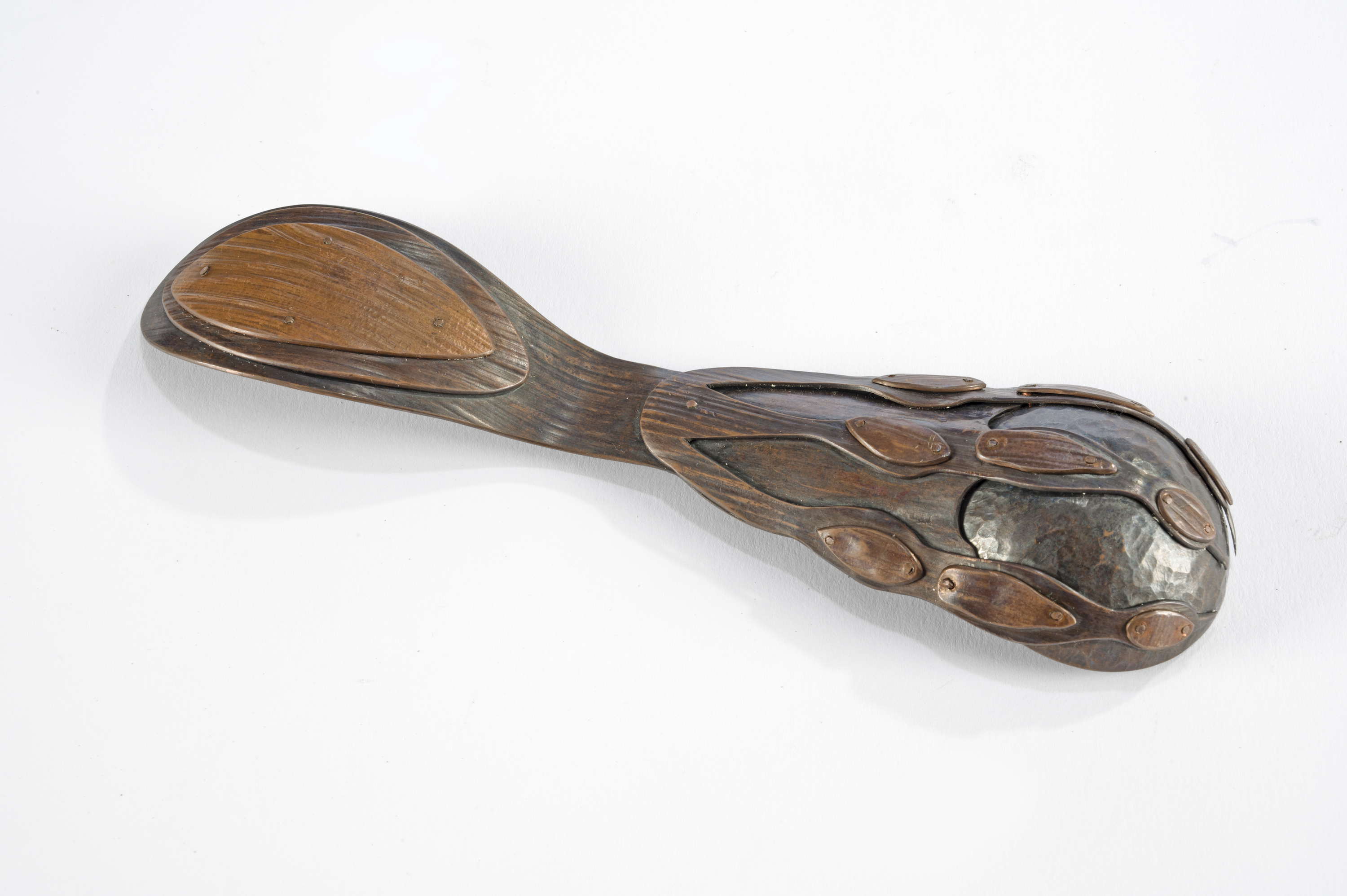 A spoon viewed from the underside made of copper with wood grain texture. Sheets of copper are cut, formed, stacked, and riveted together to look like a claw-and-ball chair foot