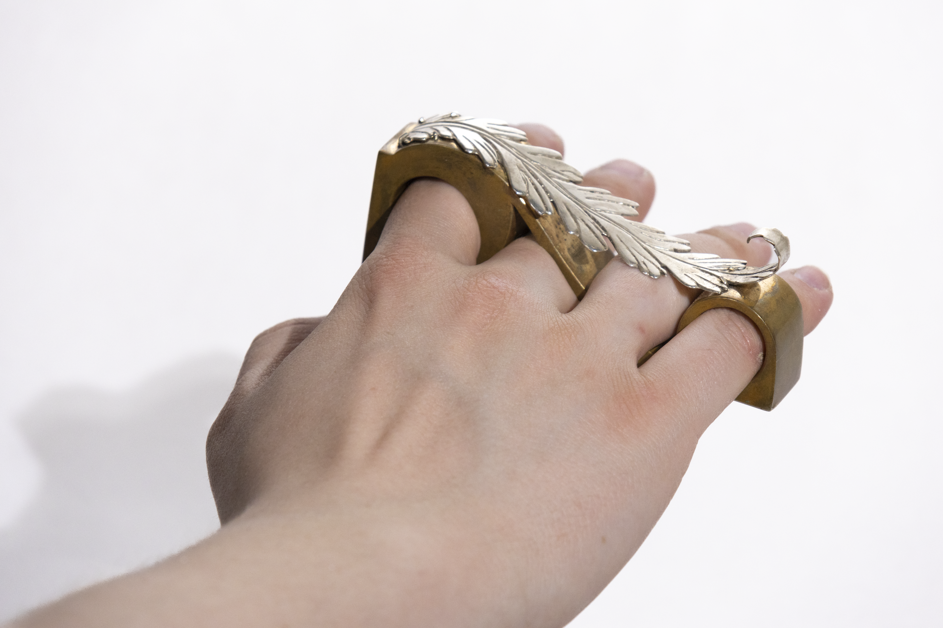 A hand reaching away from the viewer with a bronze ring that spans from the pointer to the pinky finger. The ring is capped with a curled silver acanthus leaf ornament.