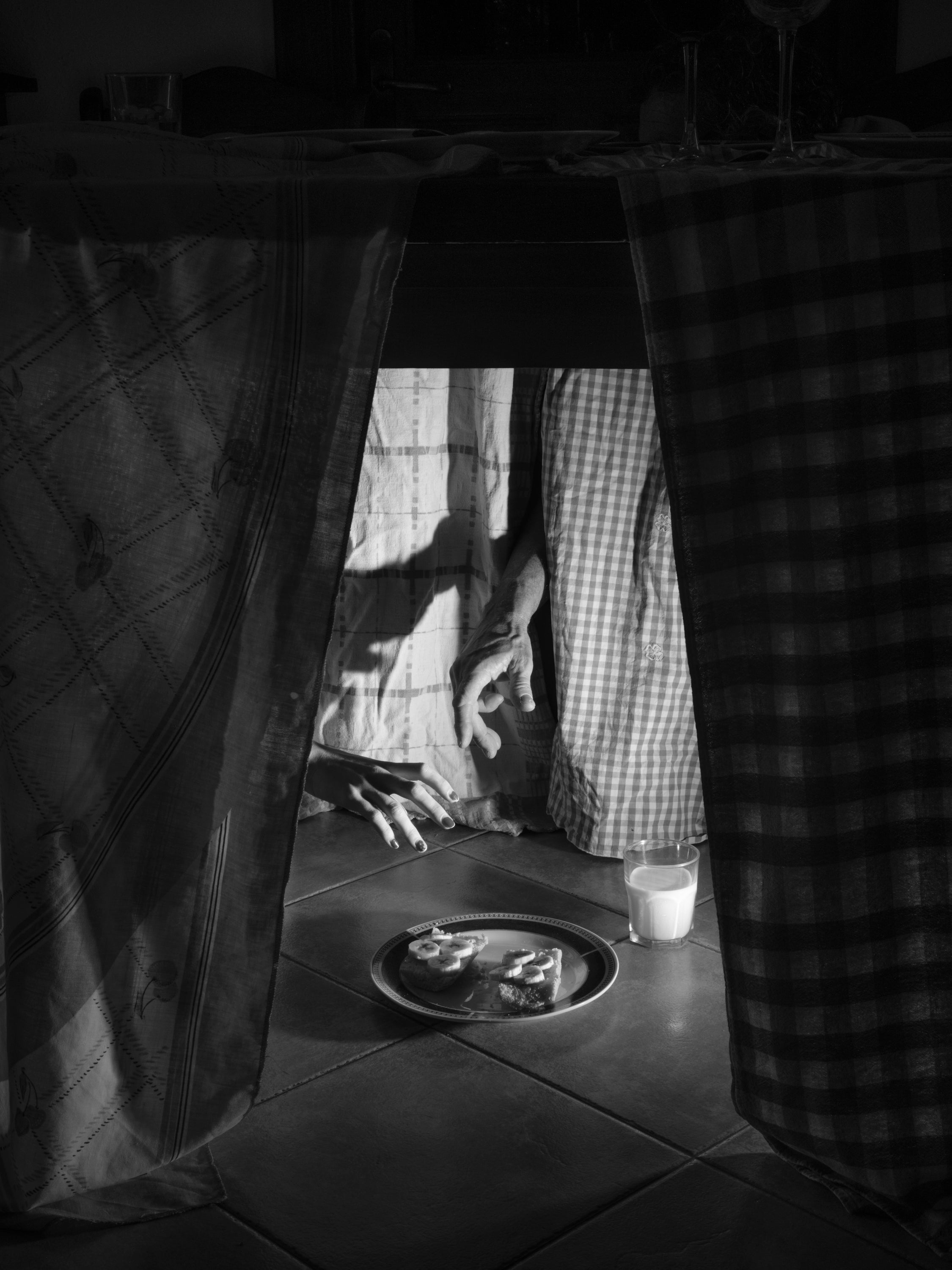 /Black%20and%20white%20photograph%20of%20the%20bottom%20of%20a%20table%2C%20with%204%20hands%20almost%20grabbing%20a%20plate%20of%20bread%20with%20banana%20and%20a%20glass%20of%20milk