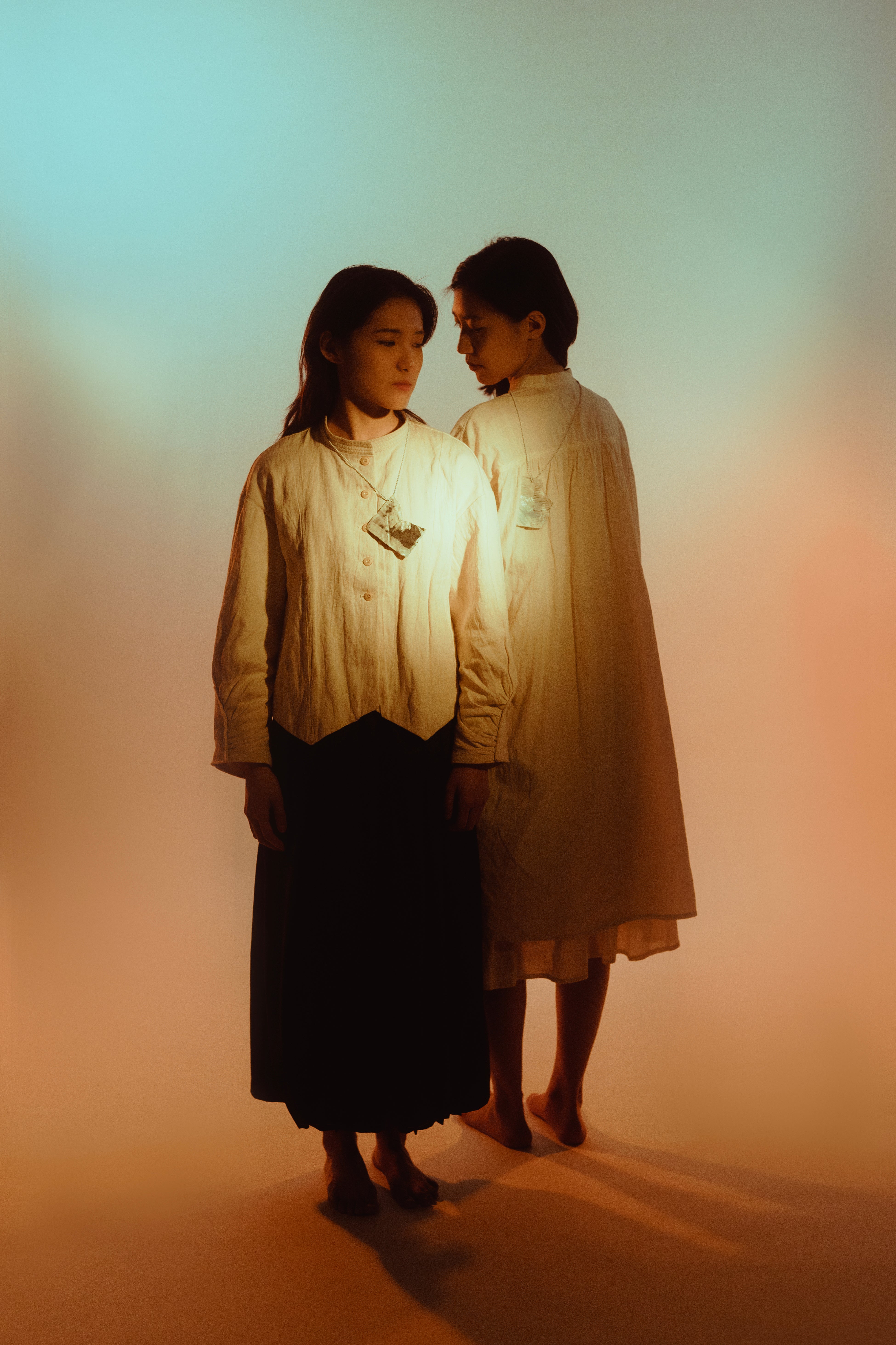 /2%20girls%20standing%20in%20the%20light%20of%20blue%20and%20orange%20mimicking%20sunrise%2C%20each%20person%20wearing%20a%20pendant.%20One%20face%20the%20front%20one%20face%20the%20back%2C%20looking%20at%20different%20directions.%20