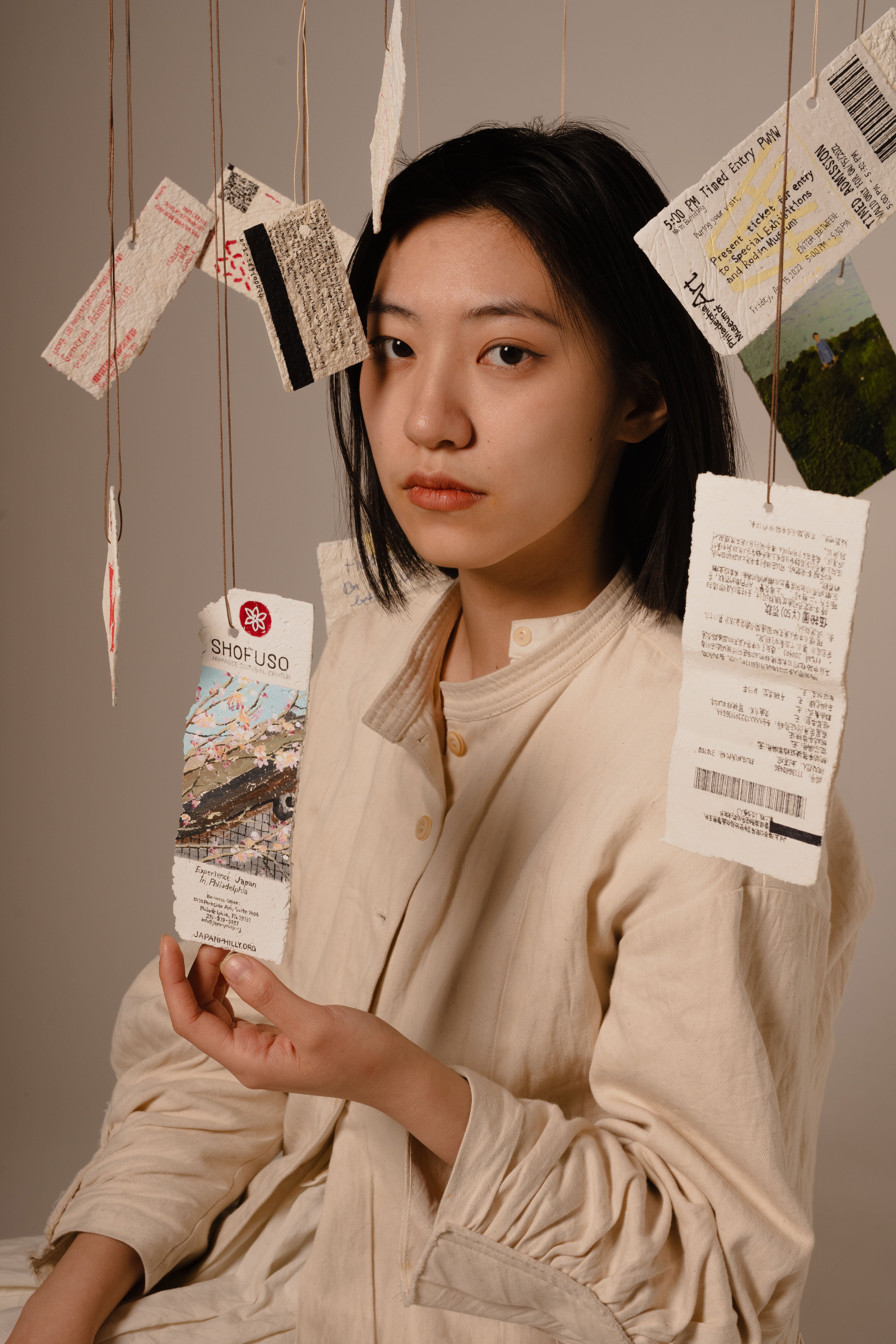 /A%20girl%20wearing%20a%20cream%20dress%20surrounded%20by%20suspended%20tickets%20painted%20on%20paper%20mache.%20Her%20one%20hand%20is%20touching%20one%20ticket%2C%20eyes%20looking%20at%20the%20camera.%20