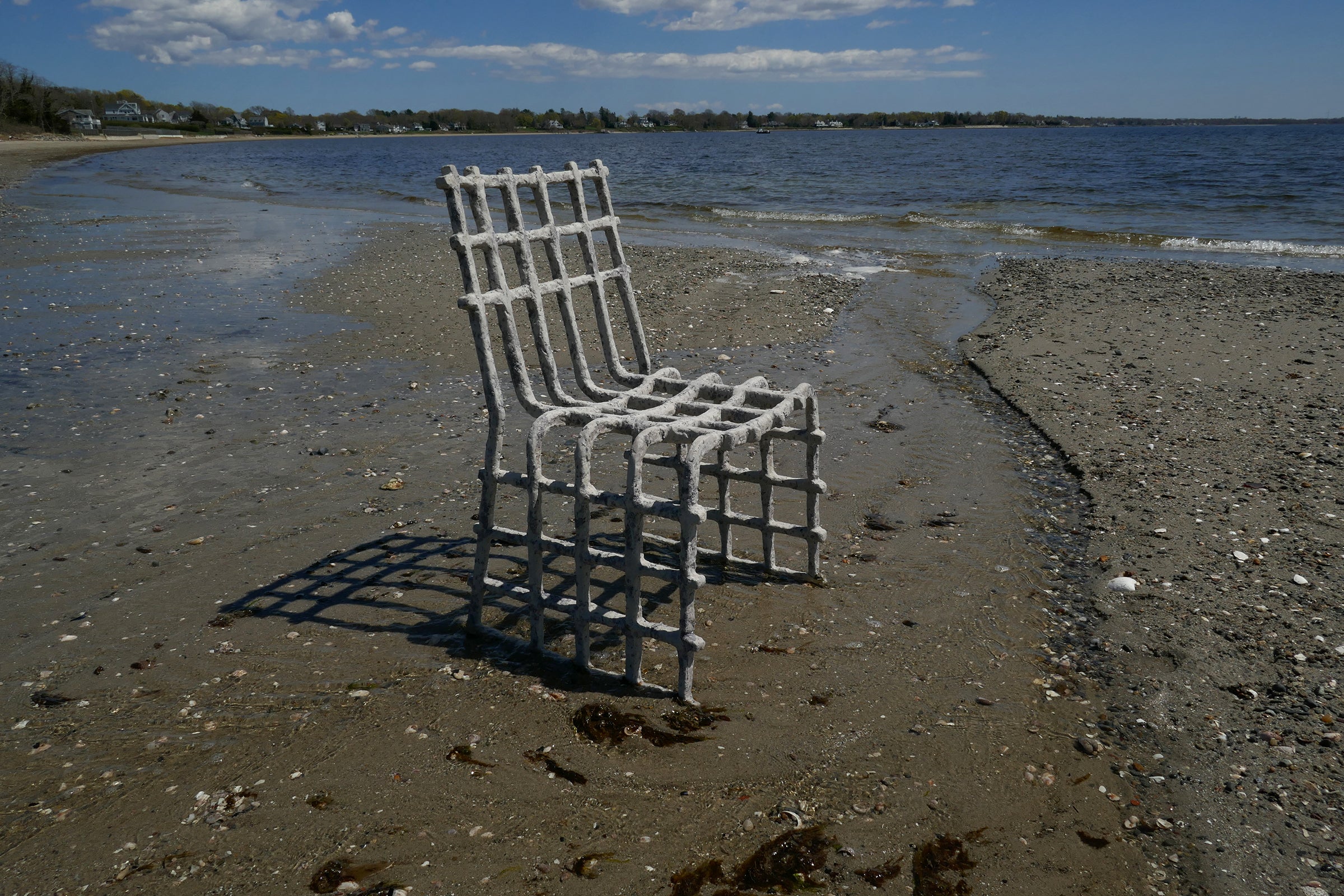 /A%20gray%20and%20blue%20oyster%20and%20mussel%20biomaterial%20chair%20on%20a%20sandy%2C%20rocky%20beach%20by%20the%20water%27s%20edge.%20Houses%20and%20trees%20are%20visible%20across%20the%20water%20under%20a%20partly%20cloudy%20sky.