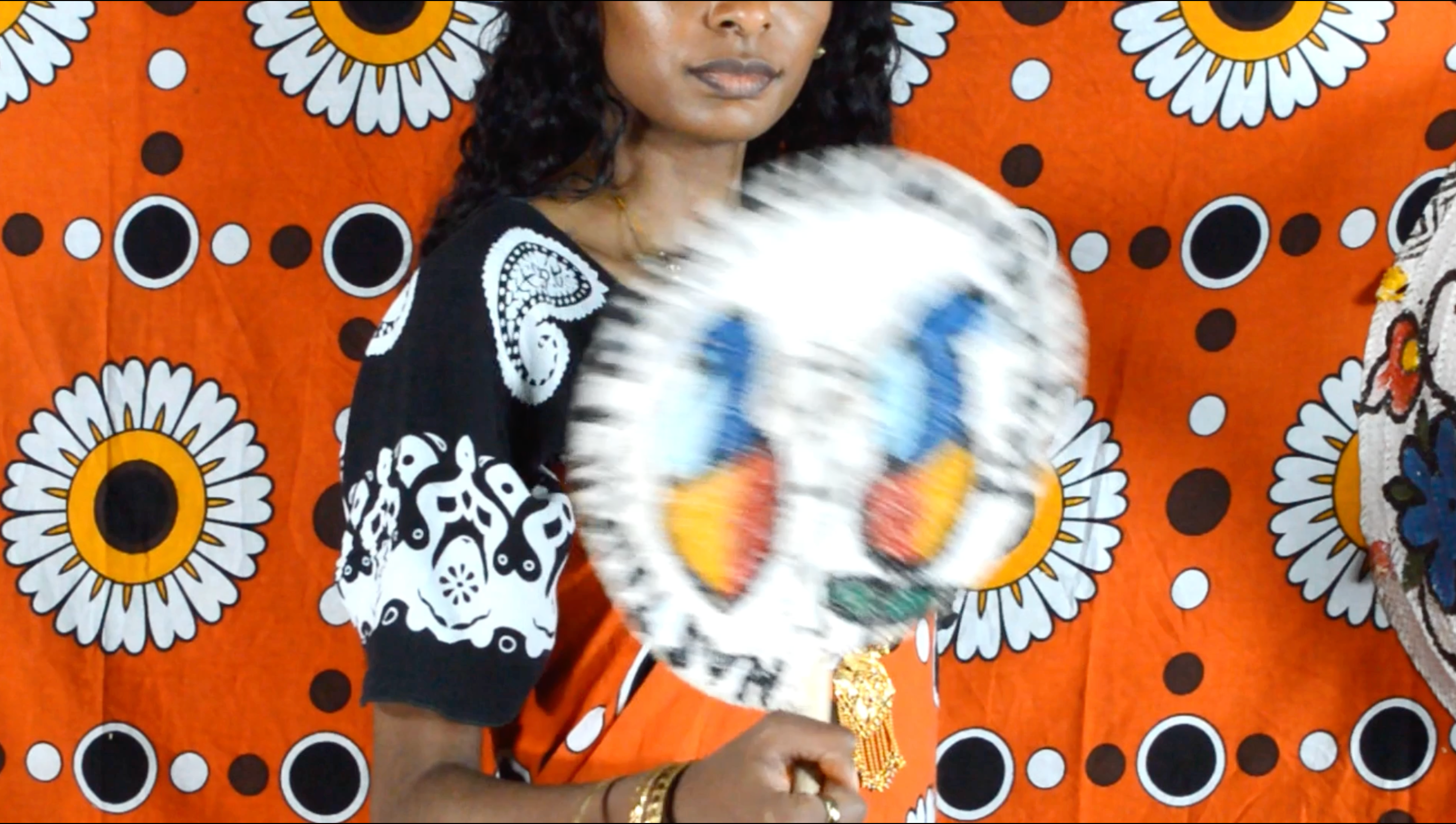 Screen shot of film from Siri Ya Mila of a woman holding a fan in front of a vibrant fabric.