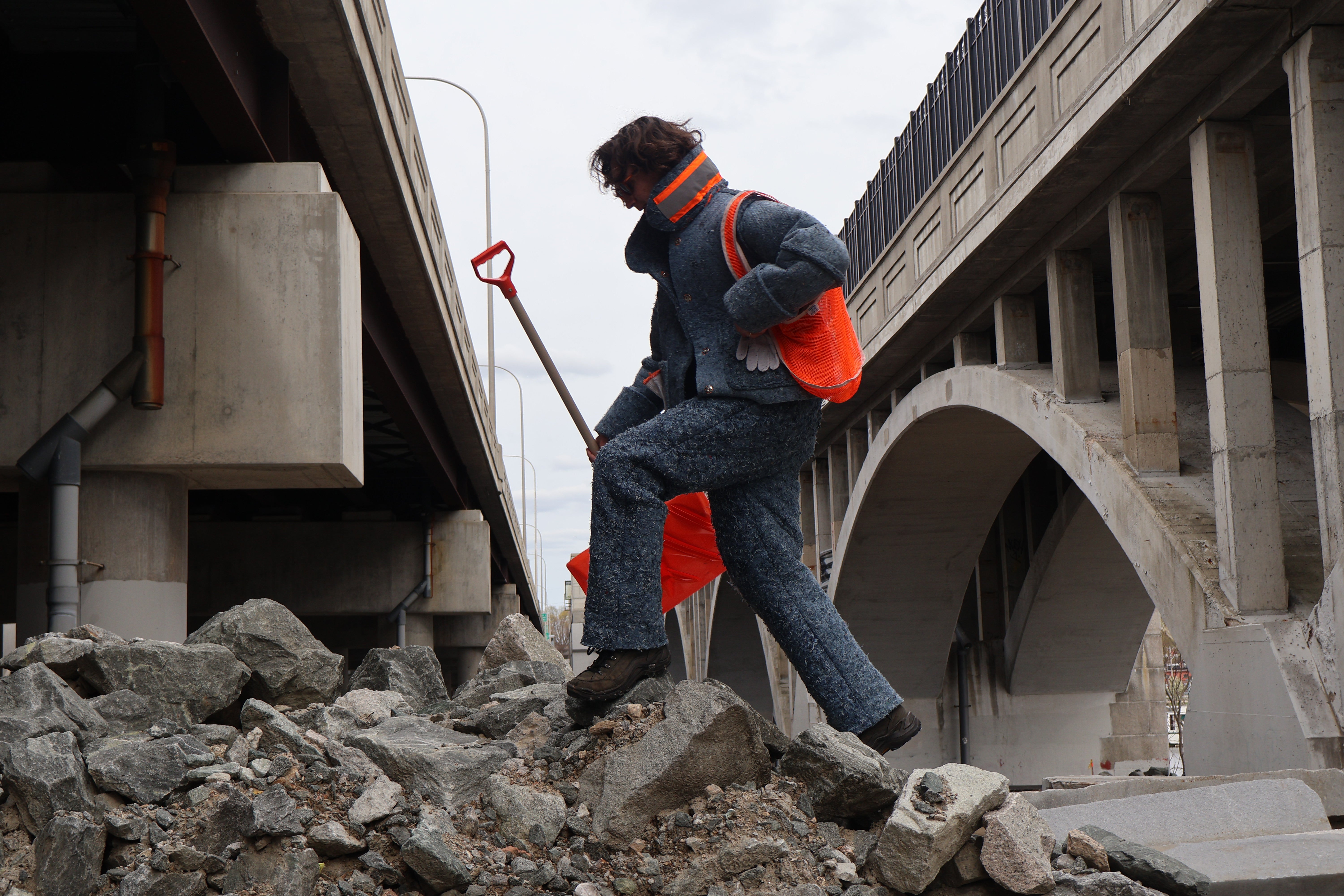 /Person%20wears%20Recycled%20Denim%20Suit%20climbing%20on%20a%20pile%20of%20construction%20rubble%20holding%20a%20shovel