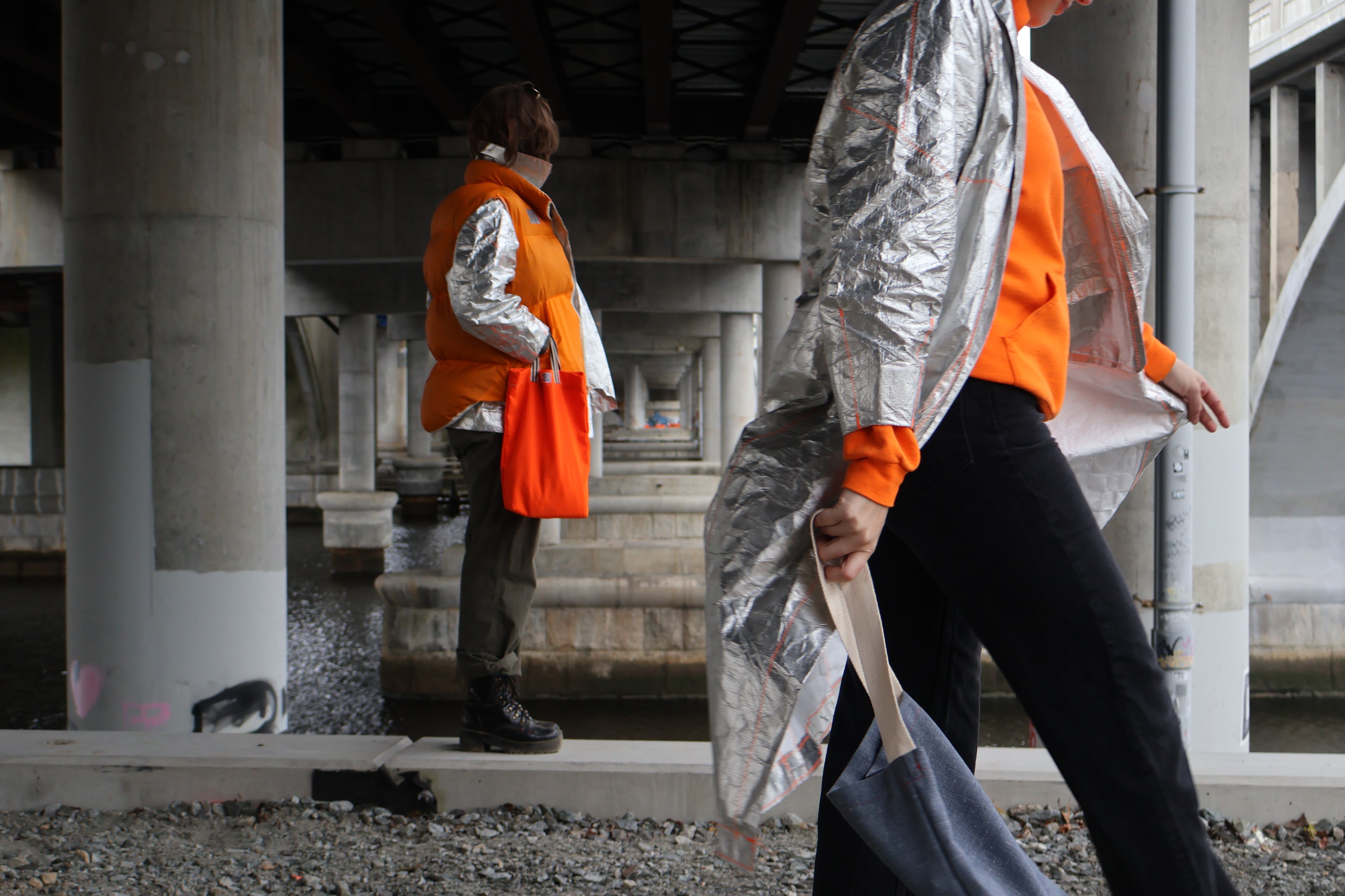 /Photograph%20of%20two%20people%20wearing%20the%20Tyvek%20Trench%20and%20Tyvek%20Coat%20layered%20with%20a%20Shoddy%20Vest%20under%20an%20overpass%2C%20holding%20bags.